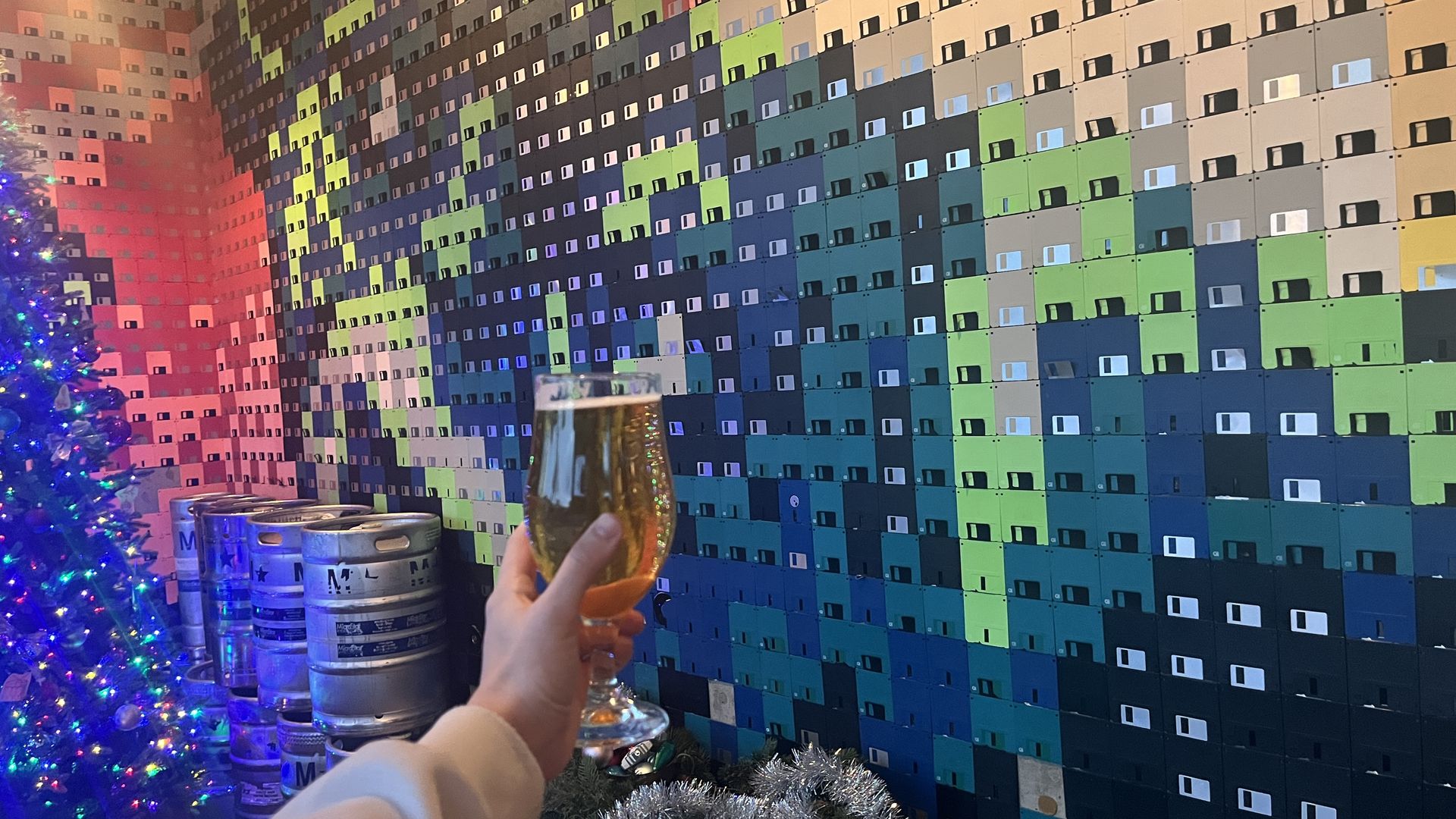 A hand holds up a beer in front of a wall mural made of floppy disks with a lit Christmas tree and kegs around.