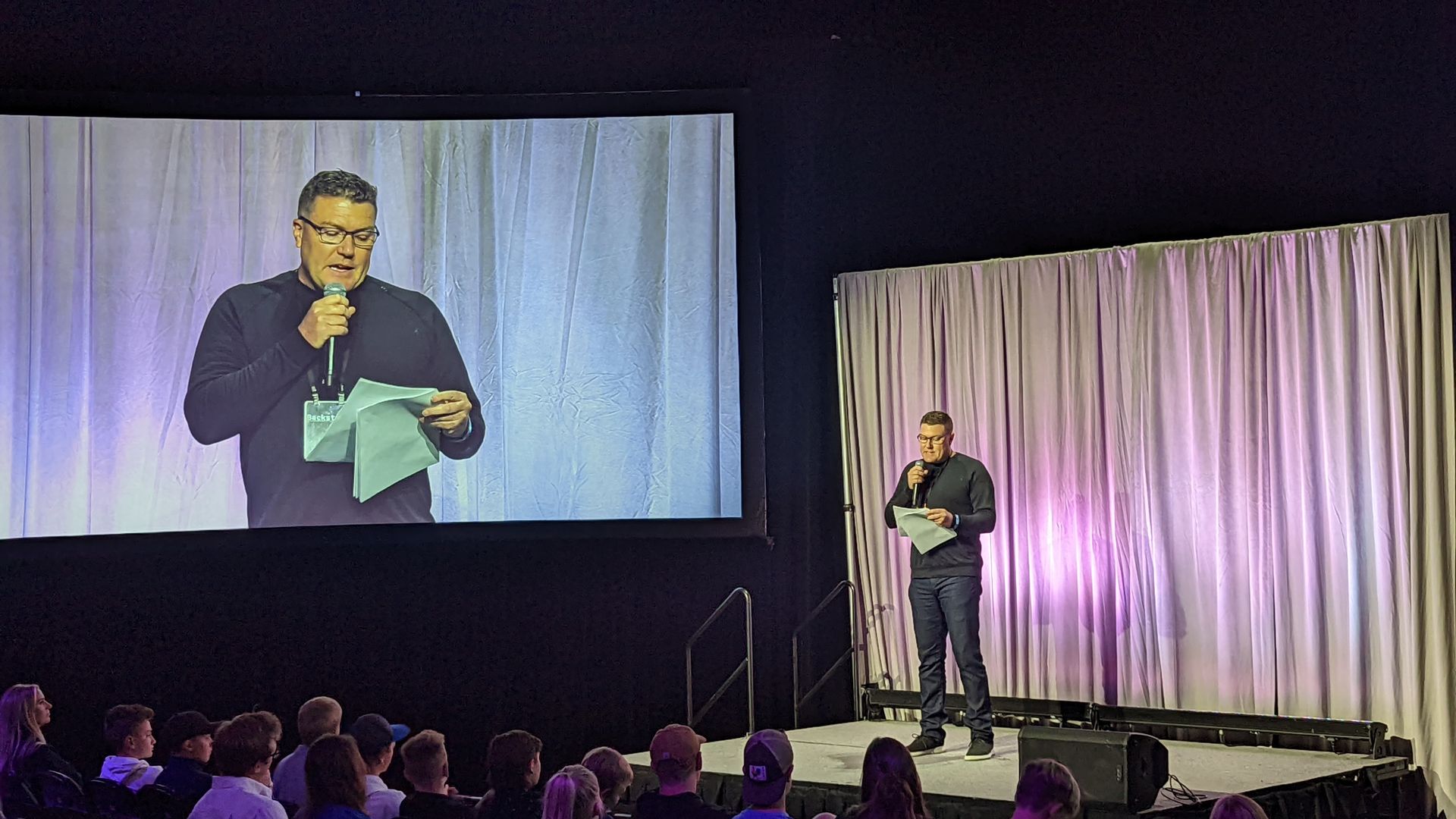 A man reads aloud onstage while a screen shows him closeup.