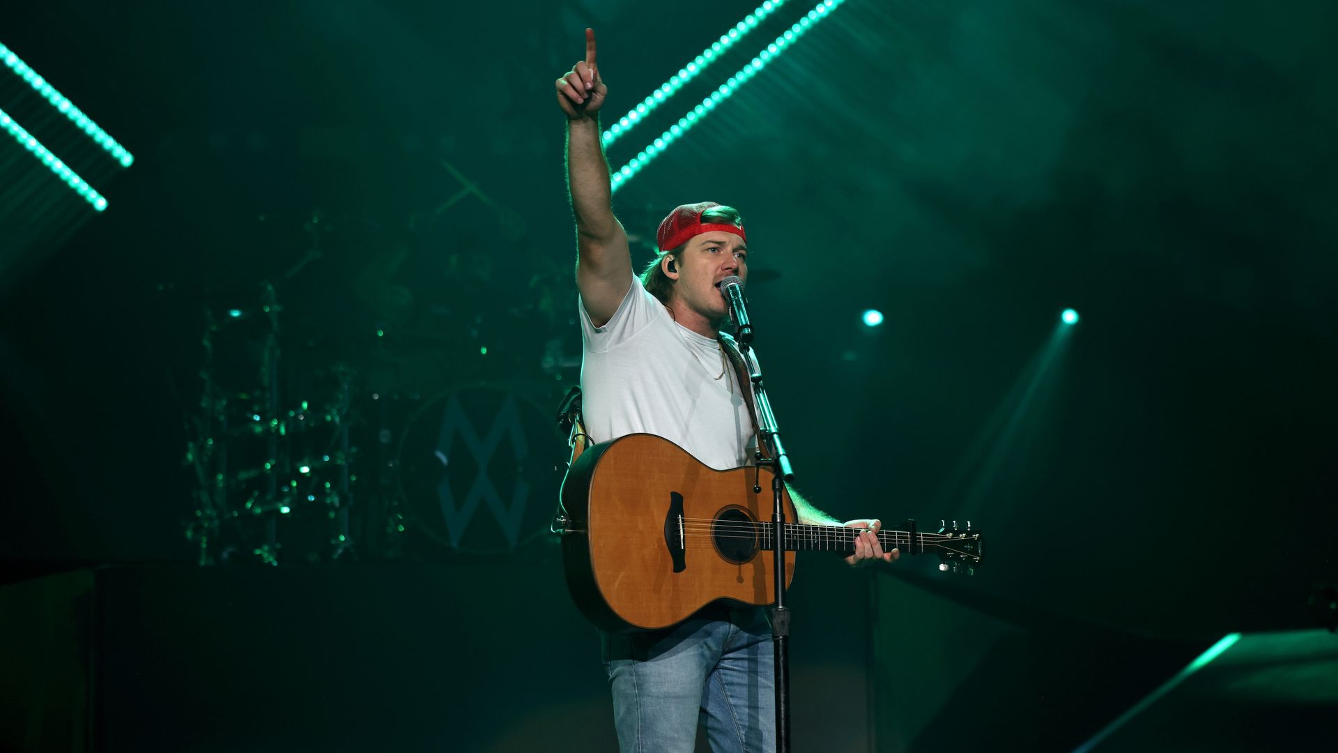 Country singer Morgan Wallen performs on stage with an acoustic guitar, holding one finger up in the air.
