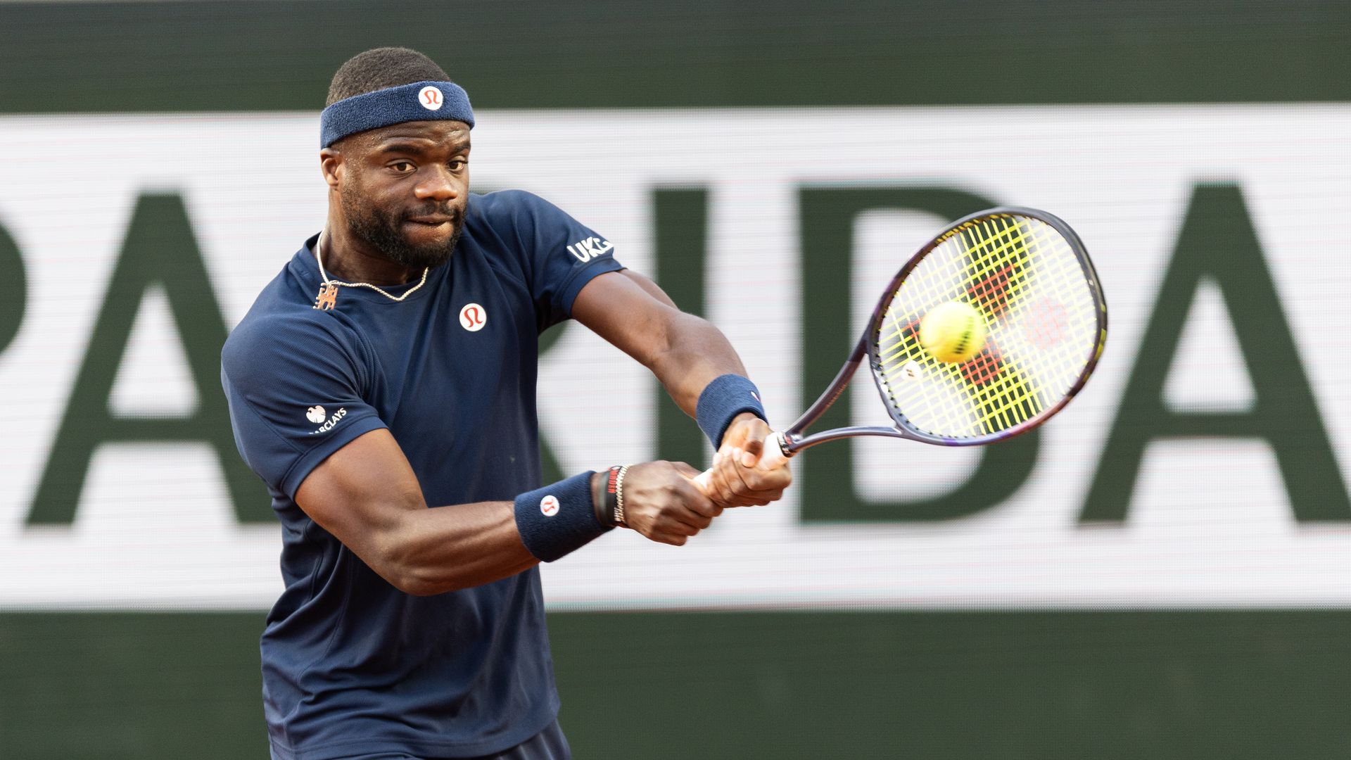 Male tennis player in navy blue outfit and blue headband swings a forehand on a tennis court; bright yellow ball near his racket, with a large blurred sponsor backdrop.