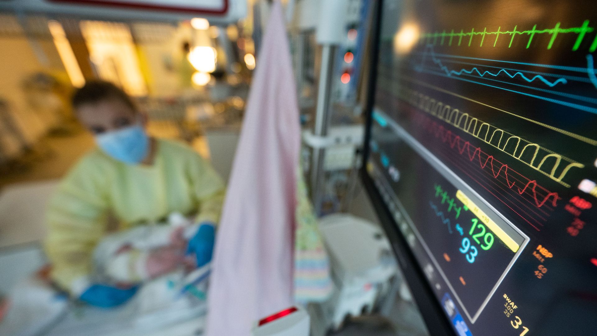 An infant in a hospital bed being taken care of by a woman in yellow scrubs and mask next to a vitals monitor. 