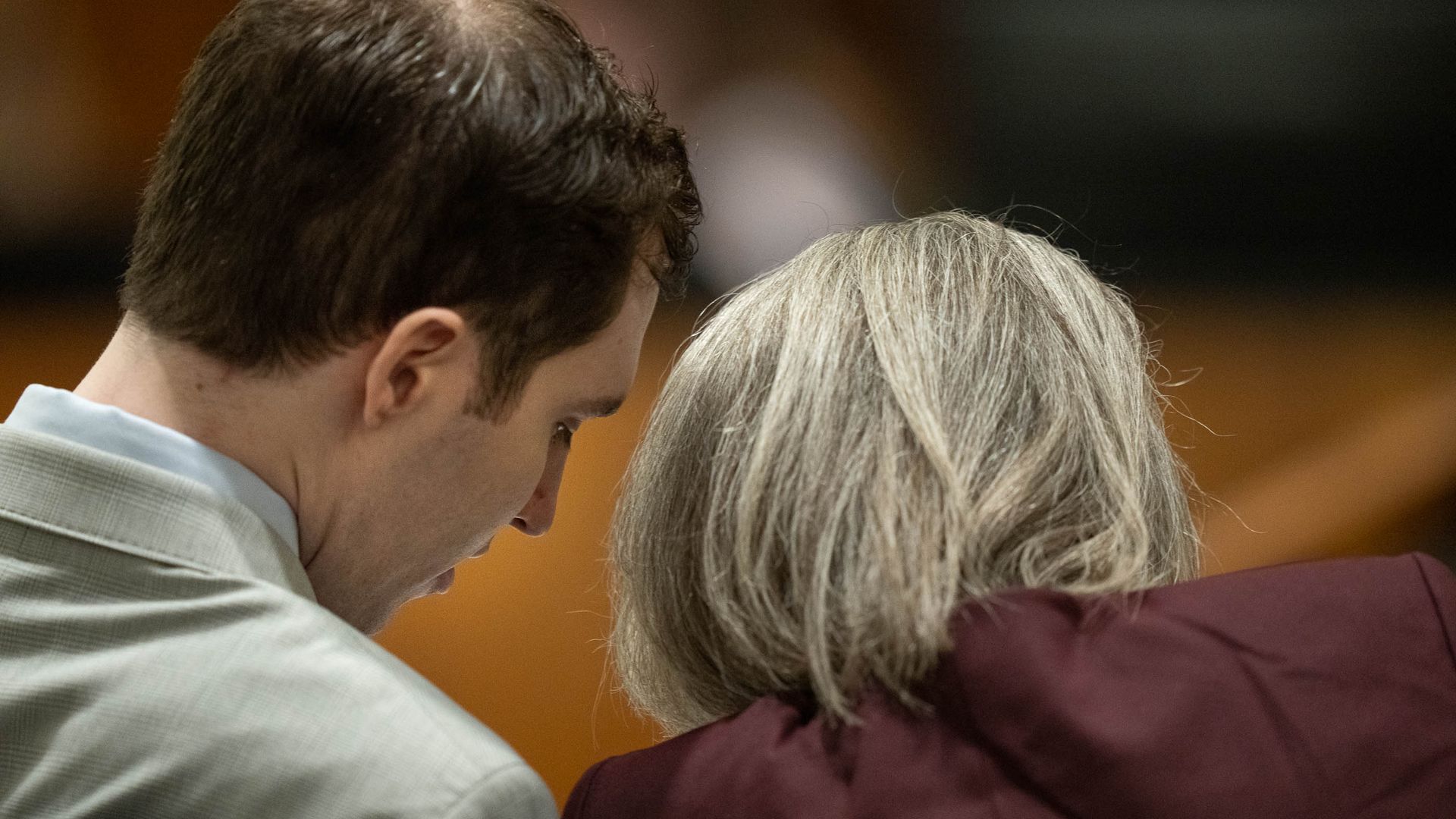 Two people sit close, heads touching in an embrace, viewed from behind: a man with dark hair in a light shirt and an older person with gray hair in a maroon top.