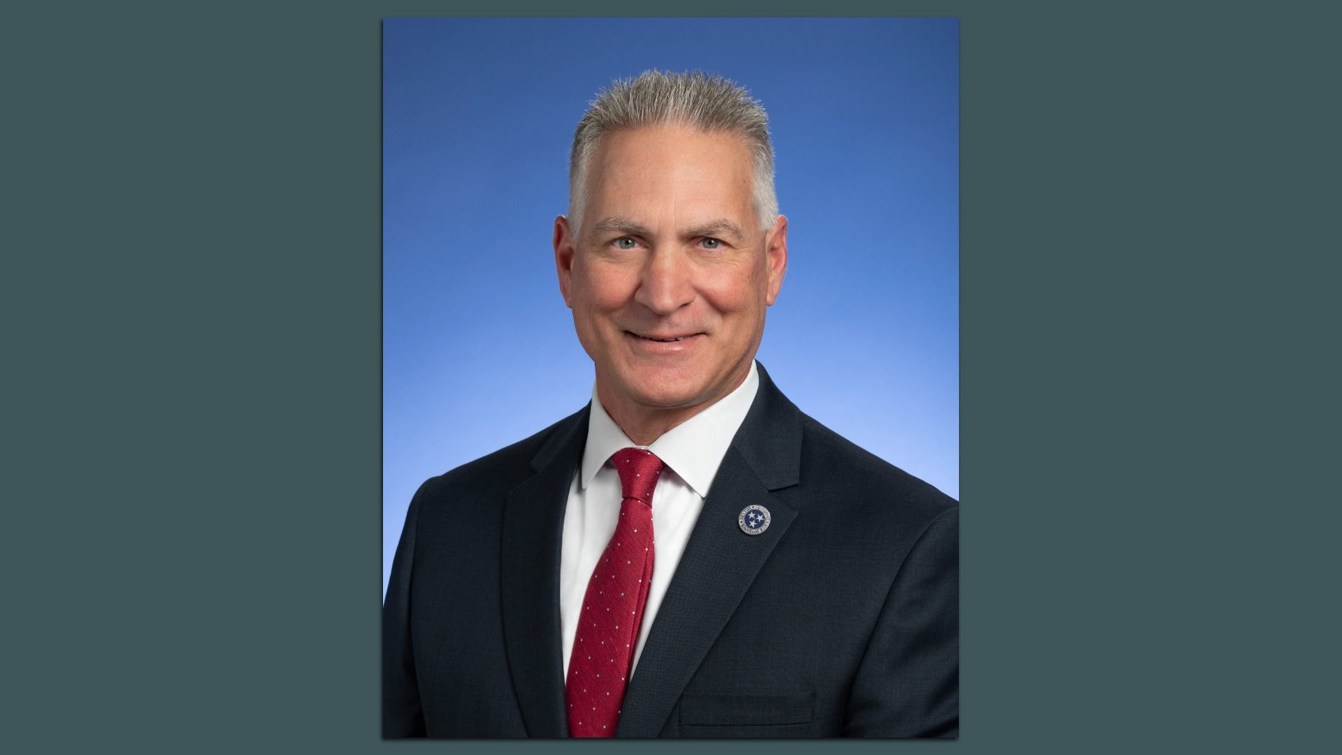 Head-and-shoulders portrait of a smiling man in a dark suit, white shirt, and red tie, wearing a circular lapel pin with a star, against a blue gradient background.