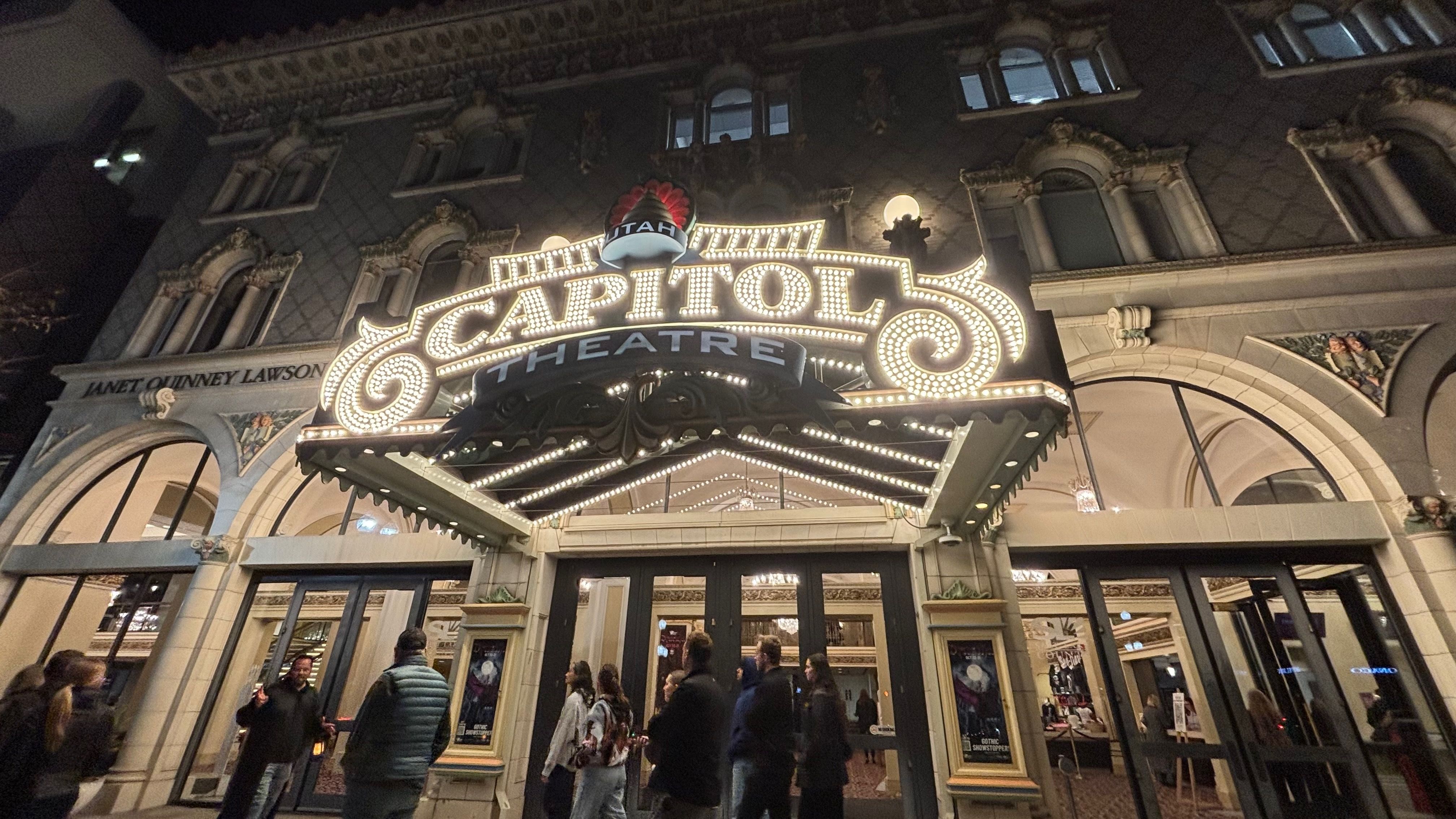 Night view of the ornate Capitol Theatre facade with a bulb-lit marquee reading "CAPITOL THEATRE" and a red crown sign above; people gather at the entrance.