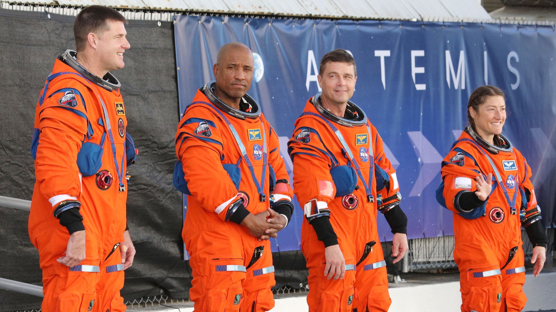Four astronauts in orange NASA spacesuits standing in front of a blue banner with the word "ARTEMIS" visible, three men and one woman, with the woman smiling and waving.