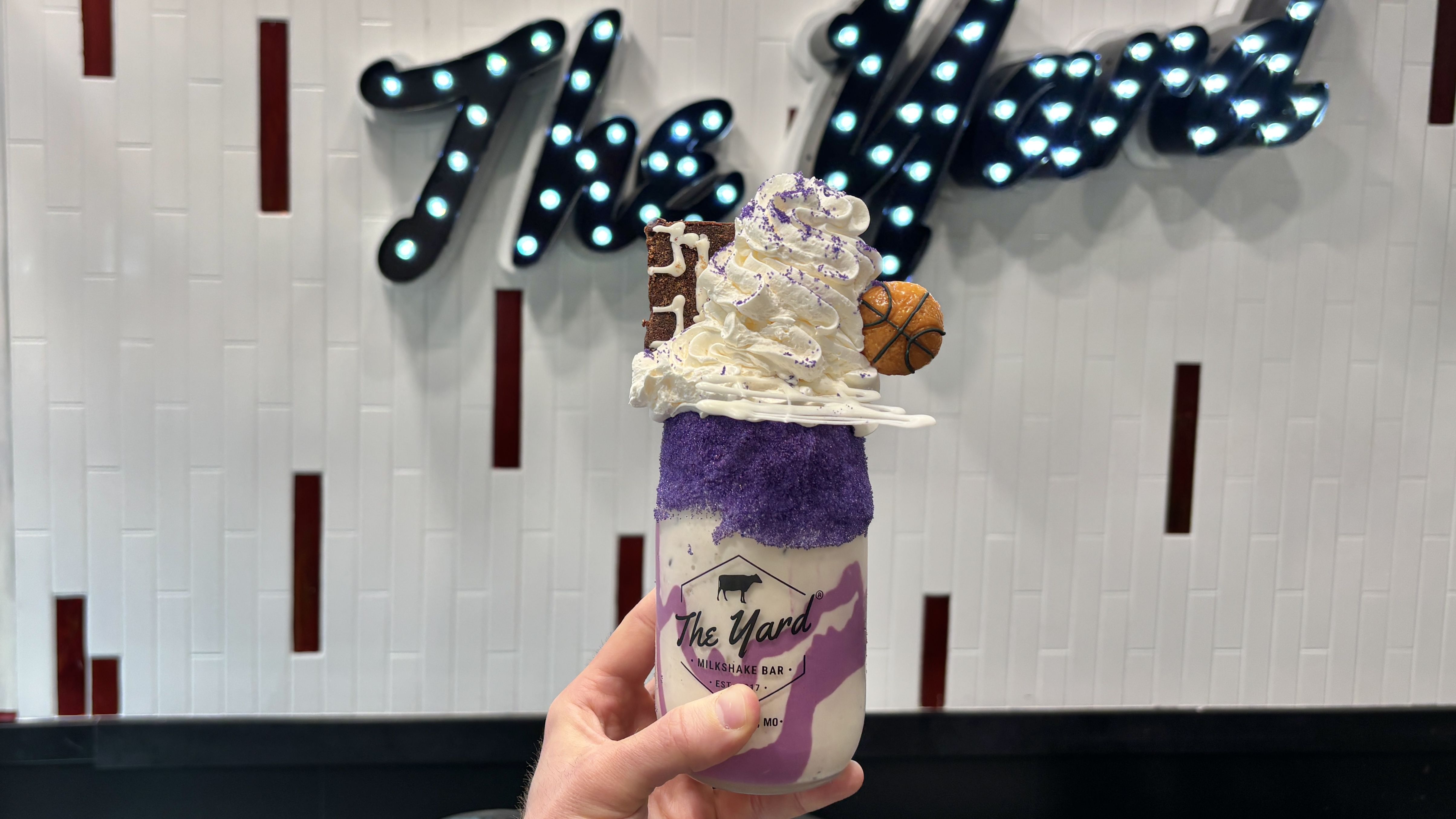 Hand holding a purple and white milkshake with whipped cream, purple sugar rim, cookie, and a small basketball decoration in front of a lighted sign that reads "The Yard" on a white tiled wall.