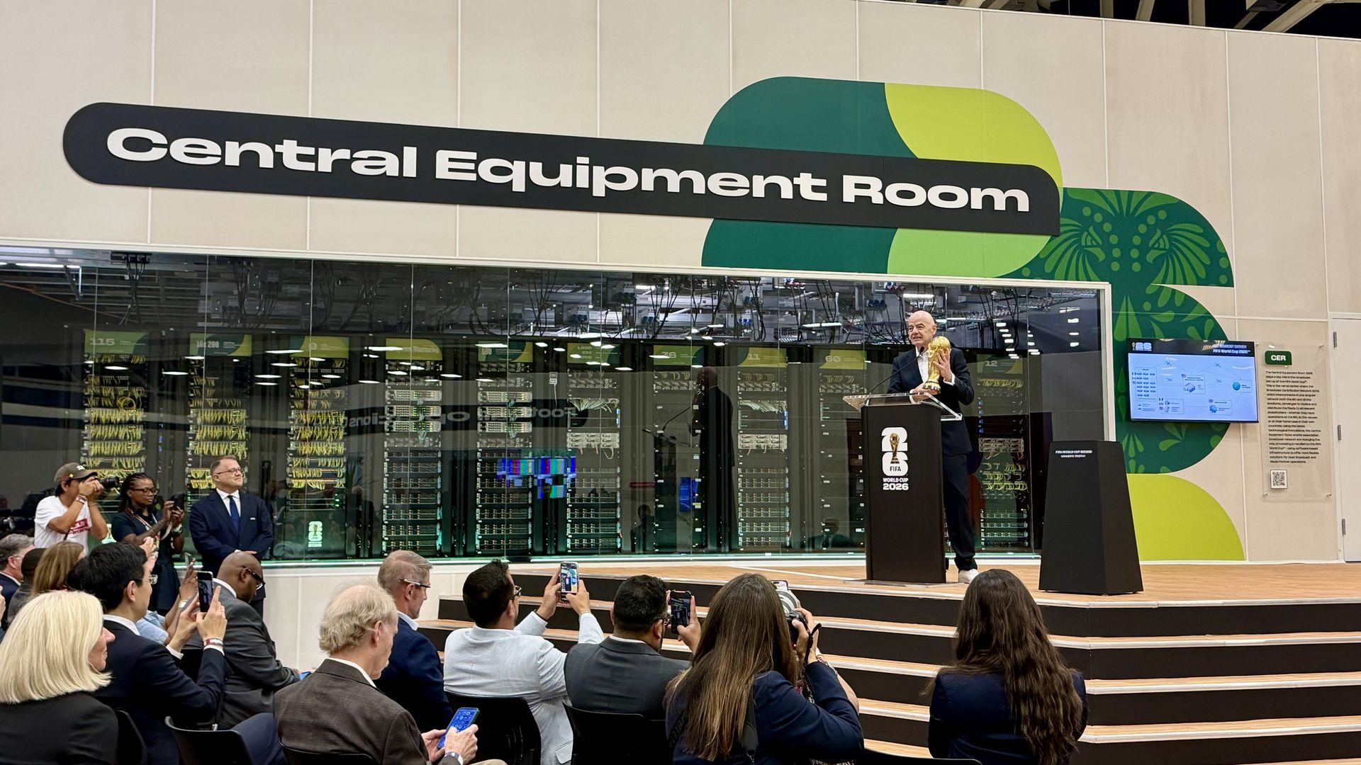 Exhibition booth with a curved black sign reading "Central Equipment Room" and green wall art. FIFA president Gianni Infantino holds a golden trophy.