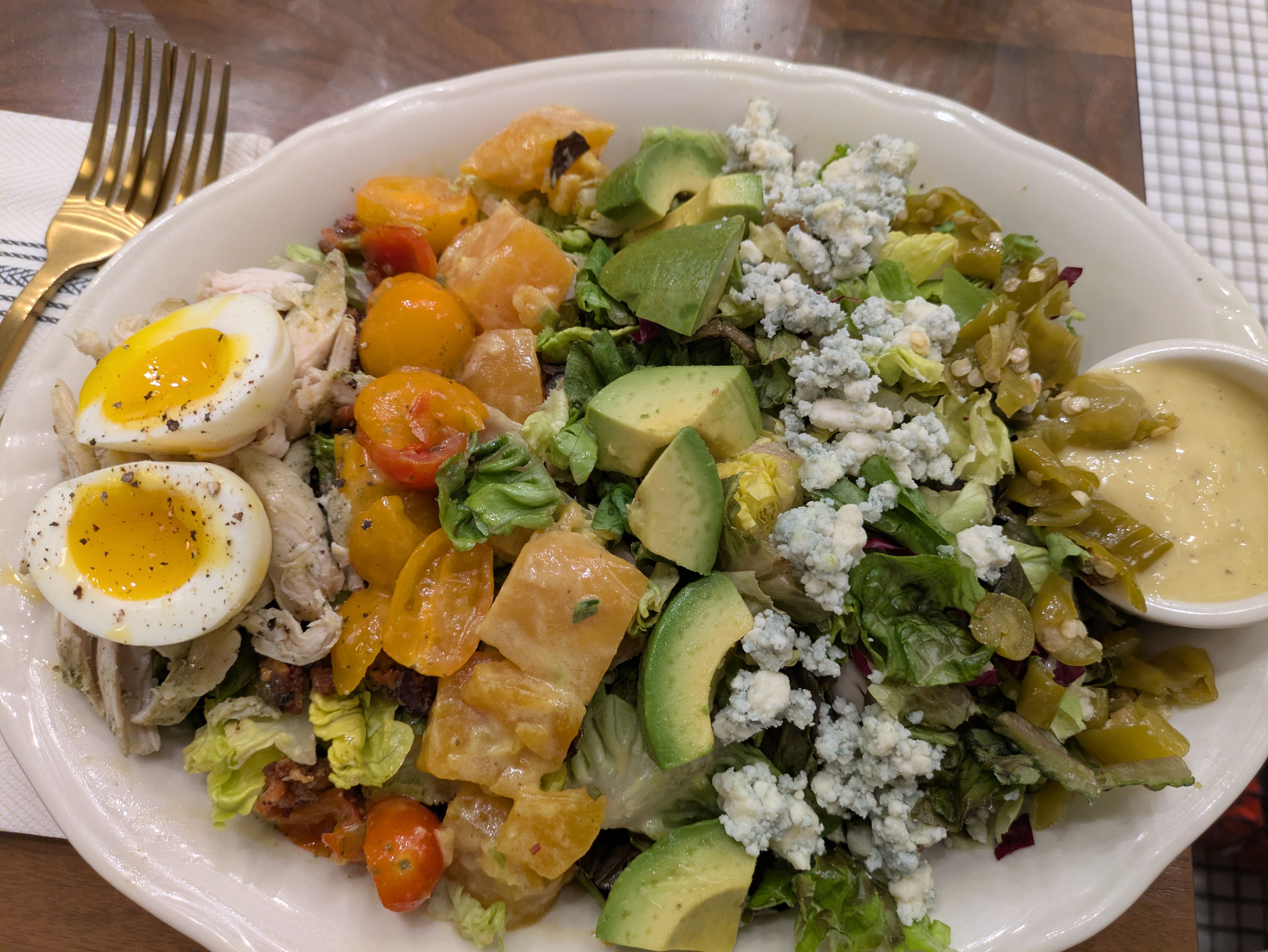A large salad bowl filled with lettuce, avocado chunks, crumbled blue cheese, halved soft-boiled eggs, orange cherry tomatoes, and a creamy yellow dressing; a gold fork rests nearby.