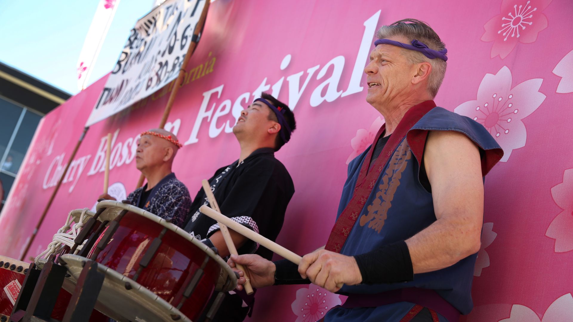 Three drummers perform on a pink Cherry Blossom Festival backdrop, wearing headbands and tunics, playing large red taiko drums.
