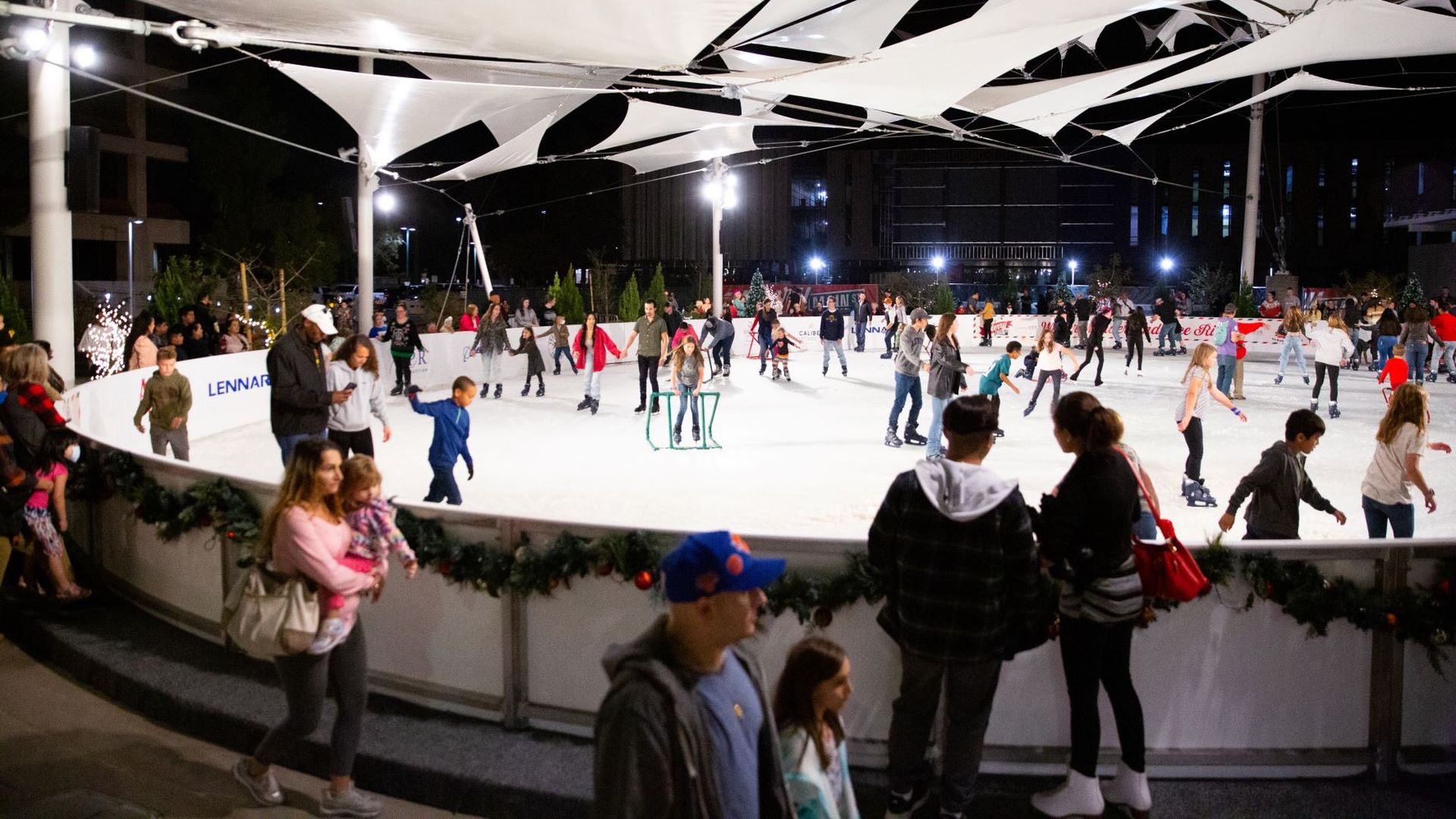 An outdoor ice rink covered by a sun shade with people walking by.