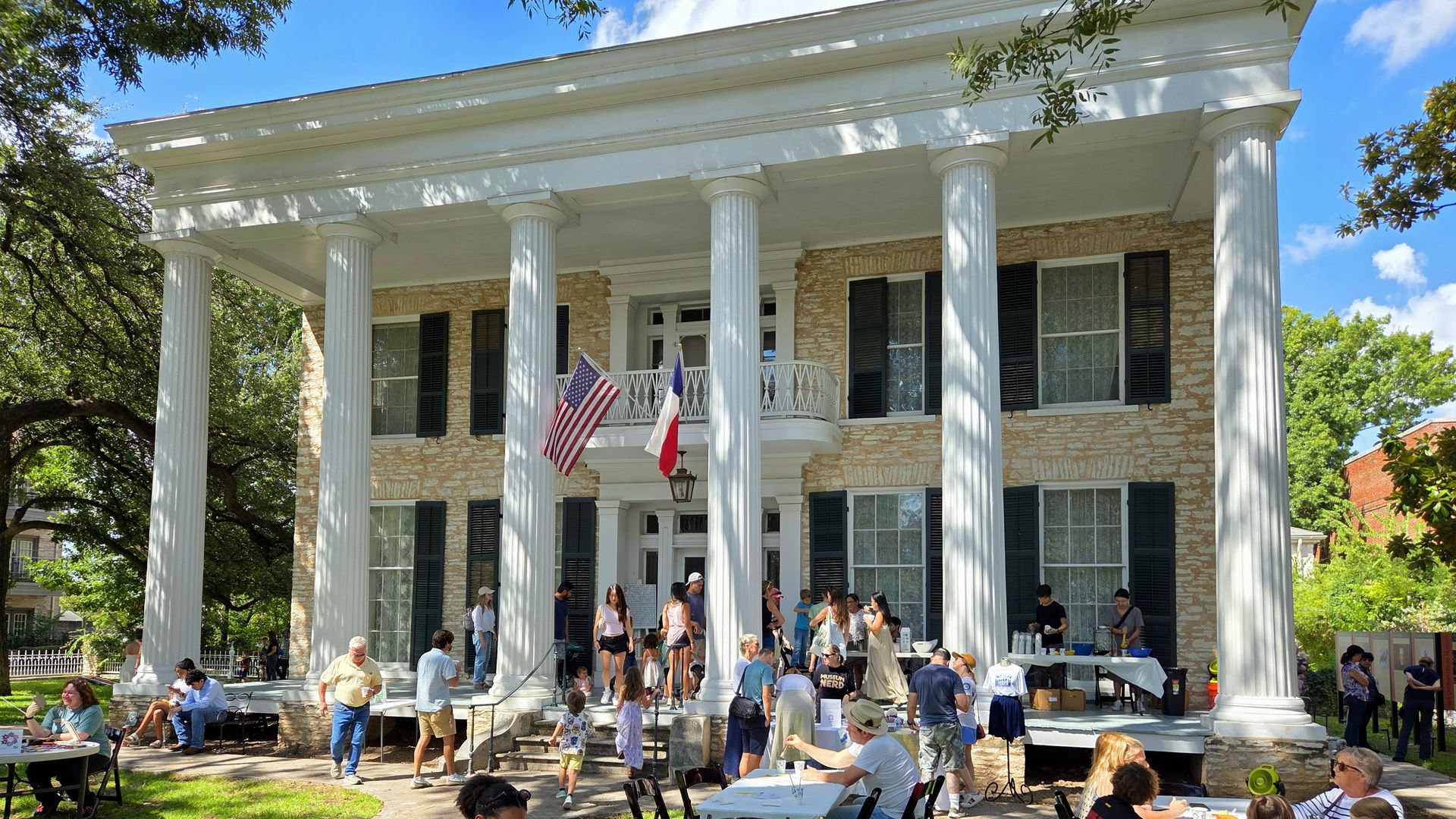 White neoclassical building with tall columns and a stone facade. American and Texas flags hang from a balcony as a crowd enjoys an outdoor craft event at tables, painting under sunny skies.