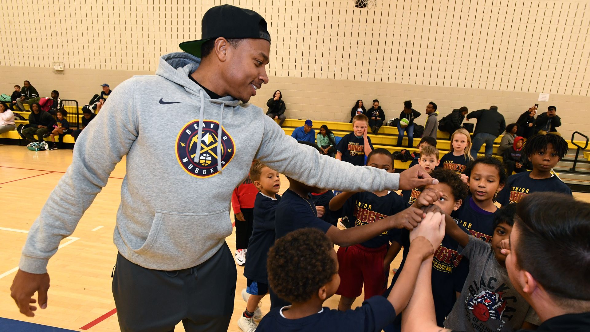 A coach in a gray hoodie and black cap high-fives a group of young basketball players in a gym, with yellow bleachers and a basketball hoop in the background.