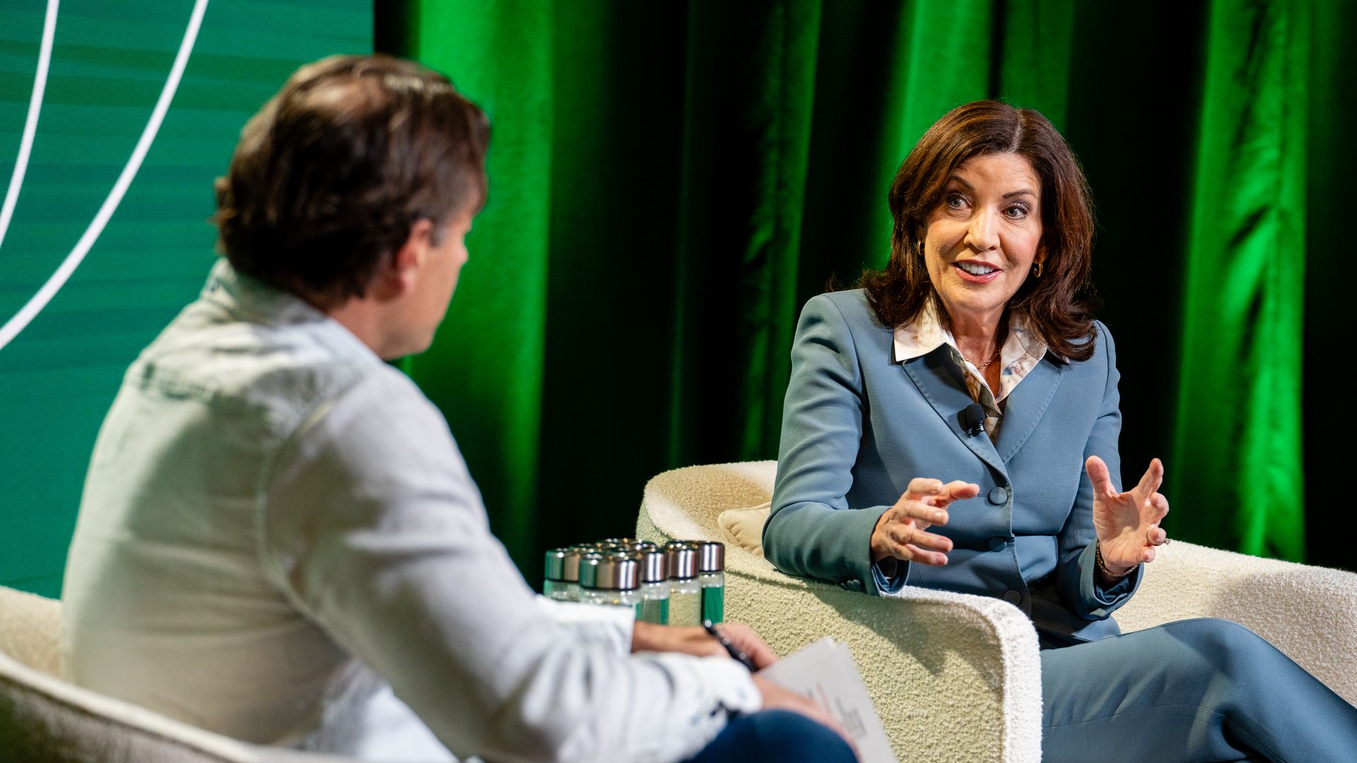 Kathy Hochul, wearing a pantsuit, sits on a chair across from Dan Primack, photographed from the back.
