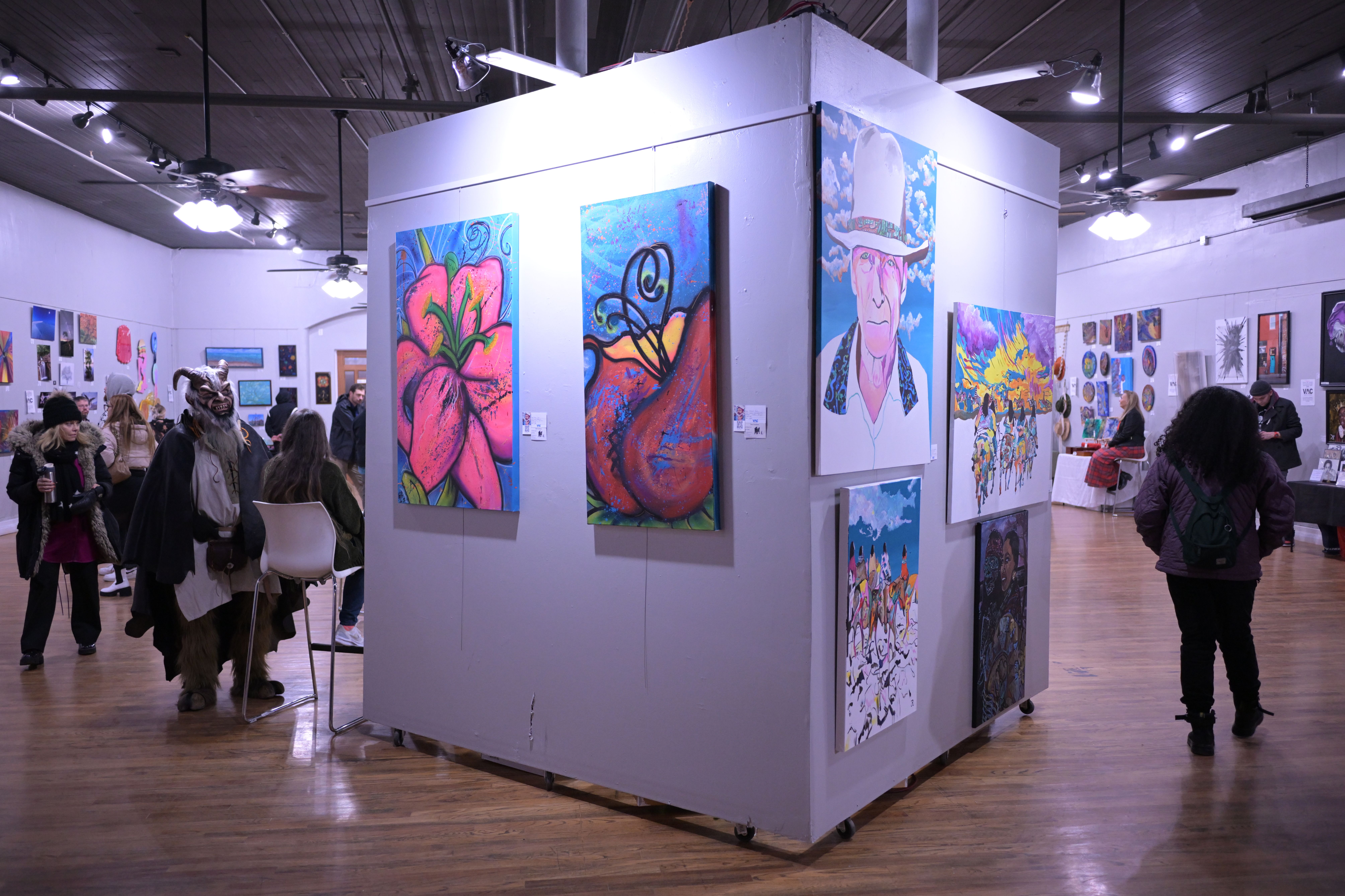A large white box inside a showroom showcases several pieces of artwork, including flowers and one depicting an elderly man. Behind it, several people are walking and viewing other artwork.
