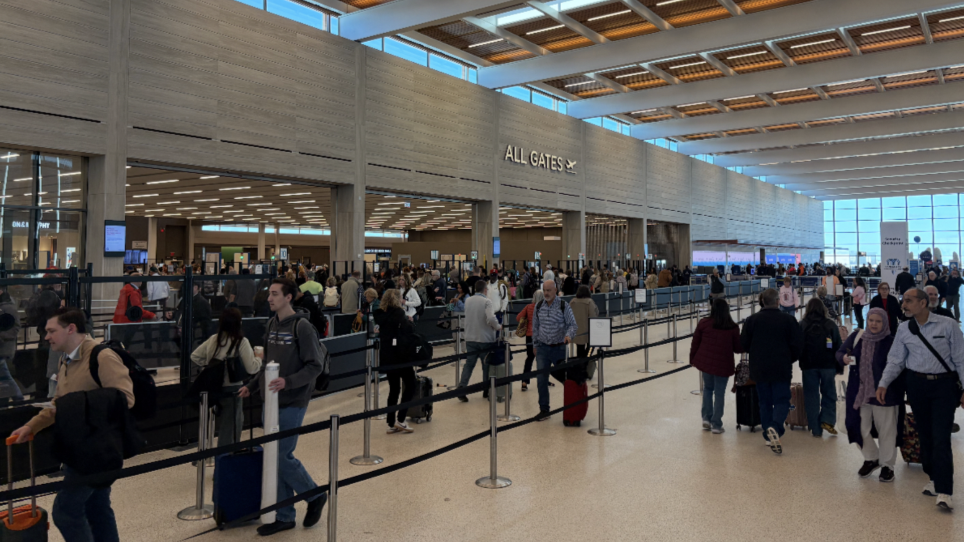 Busy airport terminal with long check-in lines and travelers with rolling suitcases; a large "ALL GATES" sign sits above the entrance as bright windows line the right.