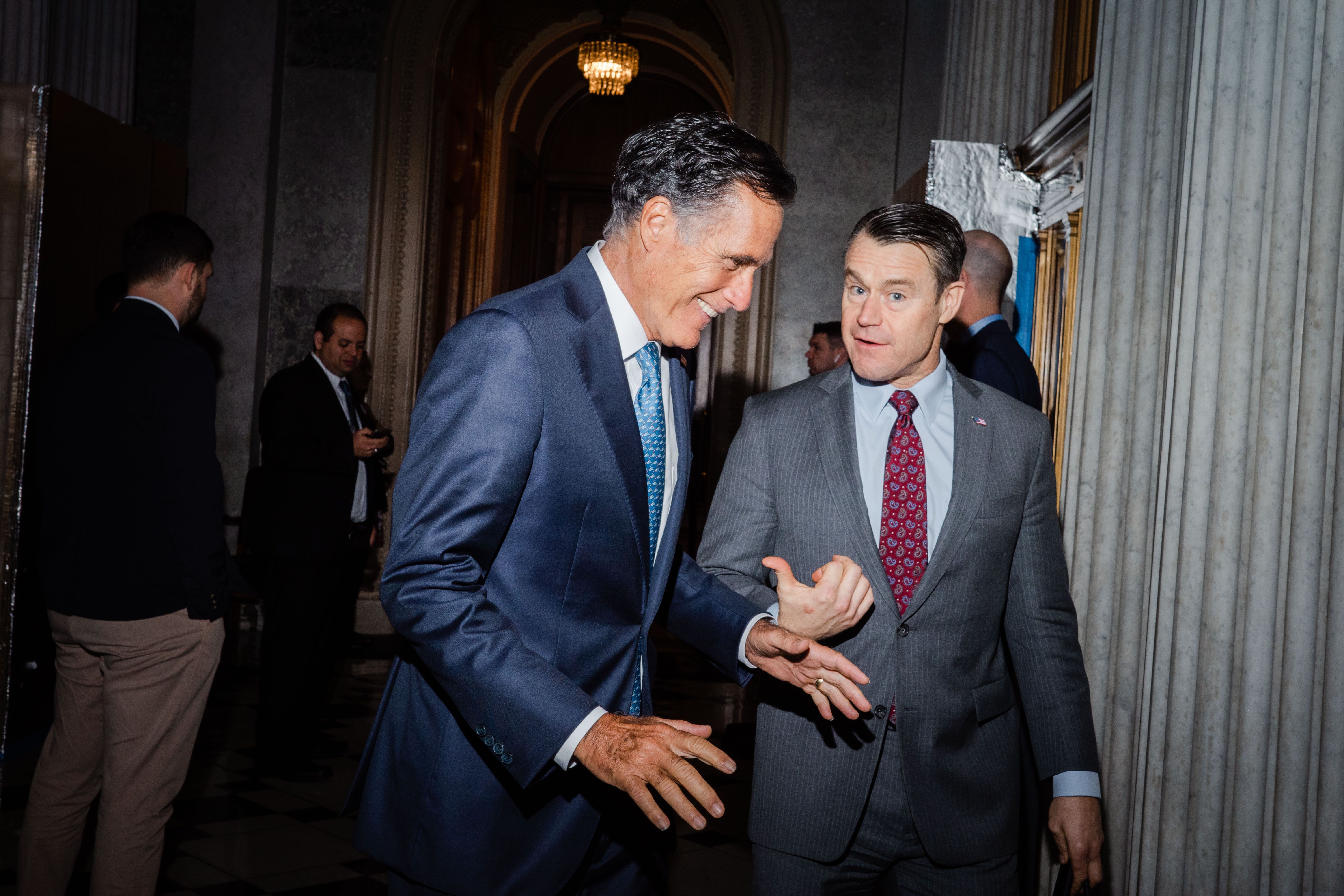 Sen. Mitt Romney laughs while Sen. Todd Young, standing to his right, smiles and points at Romney with his right thumb.
