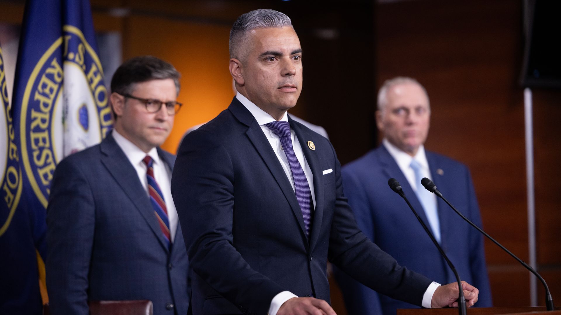 Rep. Juan Ciscomani, wearing a blue suit and flanked by Mike Johnson and Steve Scalise in a brown wooden room with House of Representatives flags.