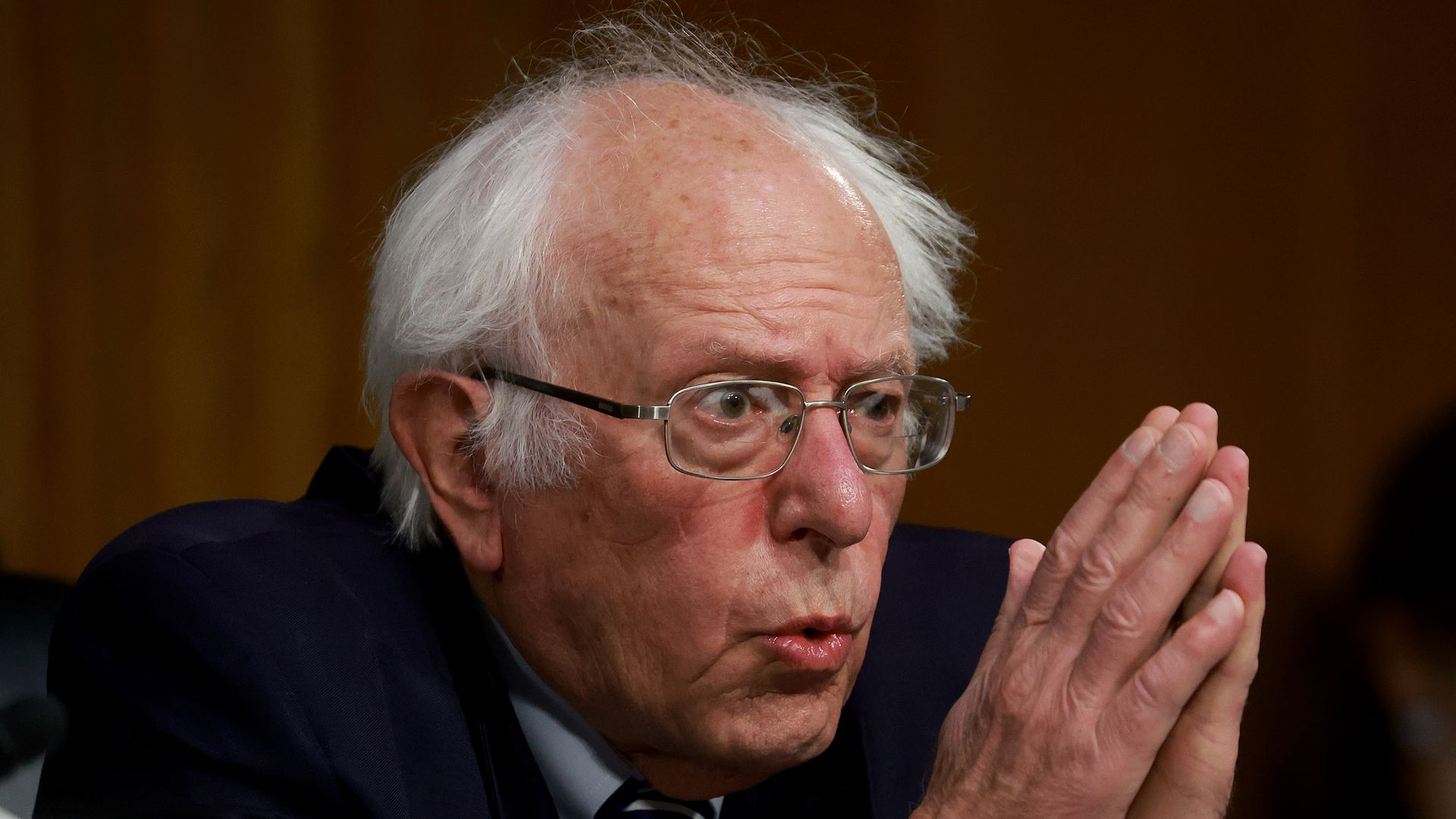 Bernie Sanders sits in a committee hearing room with his hands clasped in front of him like he's praying