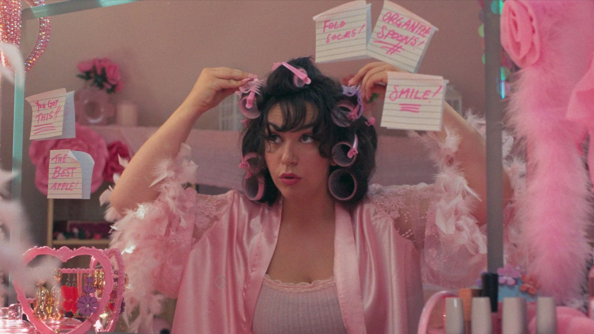 Young woman in a pink satin robe with pink curlers in her hair stands at a pink vanity mirror, surrounded by pink decor; Post-it notes read You got this and Smile!