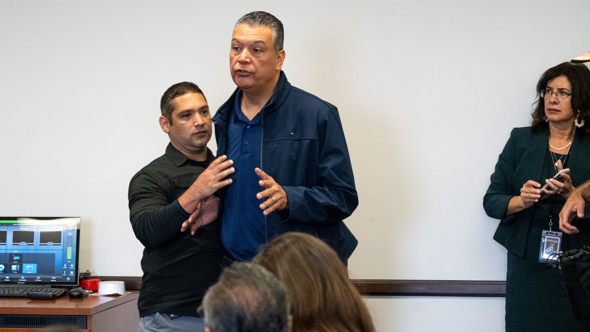 Sen. Alex Padilla, wearing a blue windbreaker, is grabbed by an FBI agent wearing a black shirt in front of a white wall.