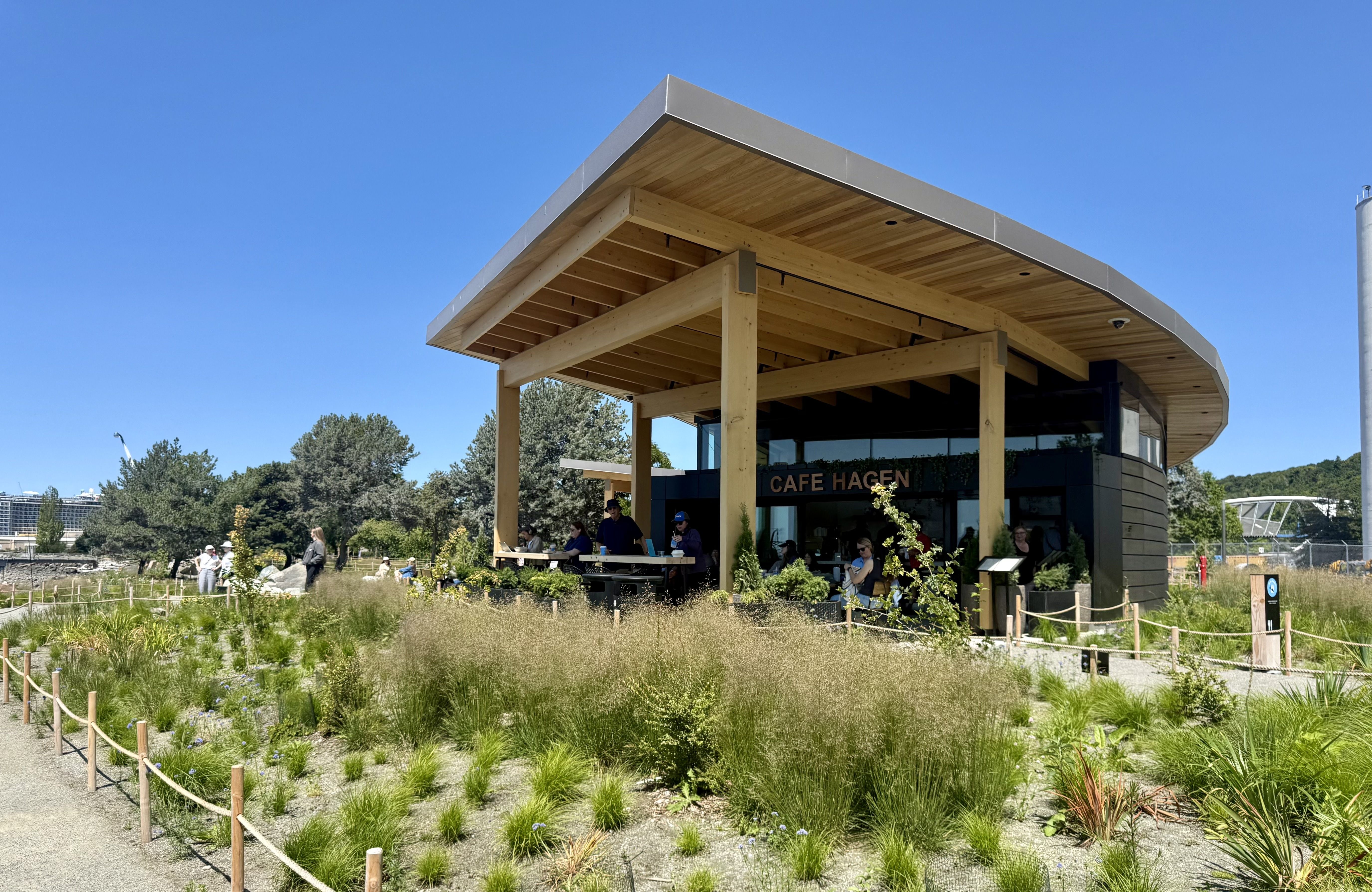 Open-air cafe under a large wooden pergola with a flat roof and exposed beams. The sign reads "CAFE HAGEN"; people sit at outdoor tables beneath blue sky and surrounding greenery.