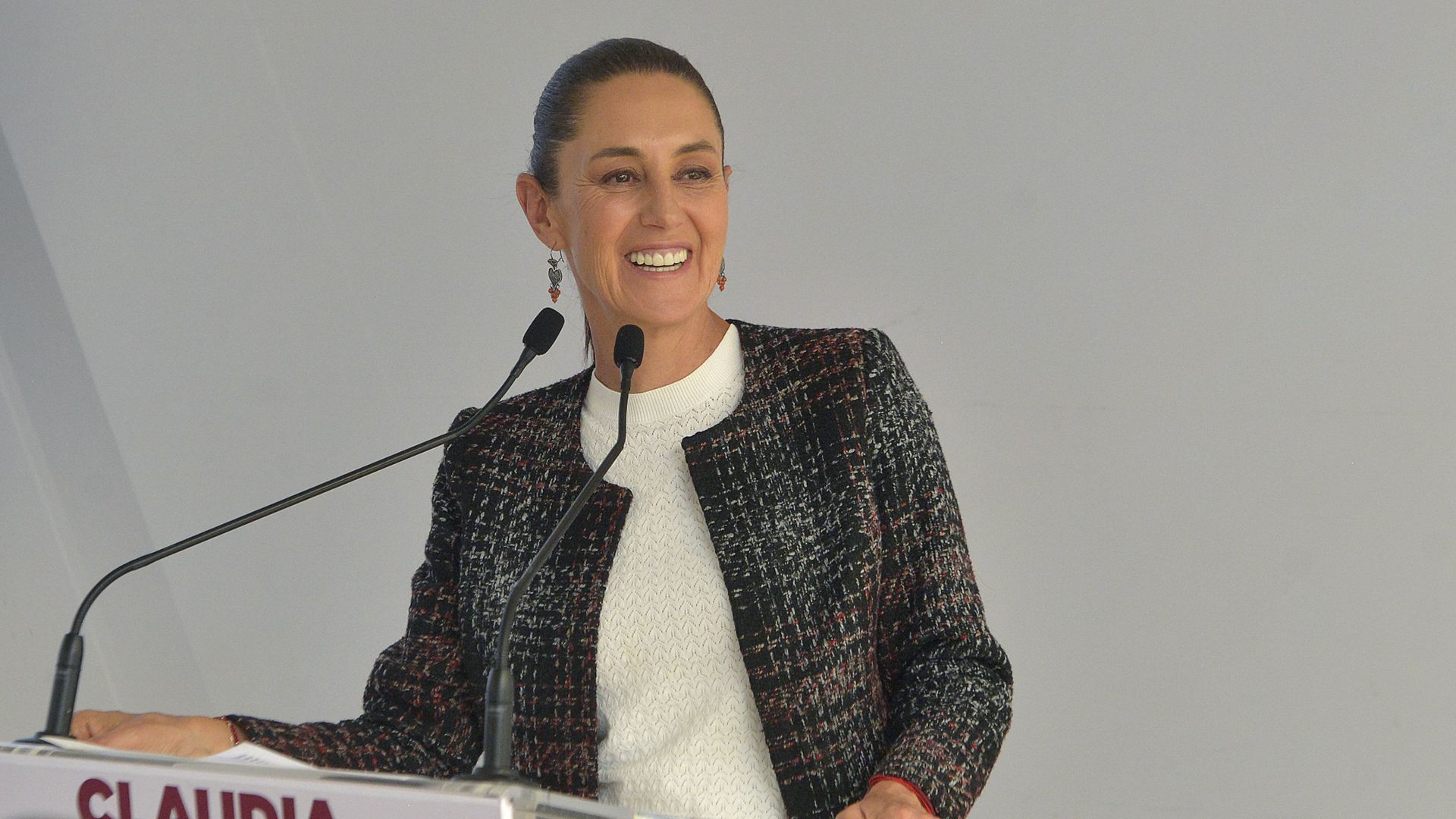Incoming Mexican President Claudia Sheinbaum stands at a podium with two small microphones in front of her.She is smiling and looking to her left. 