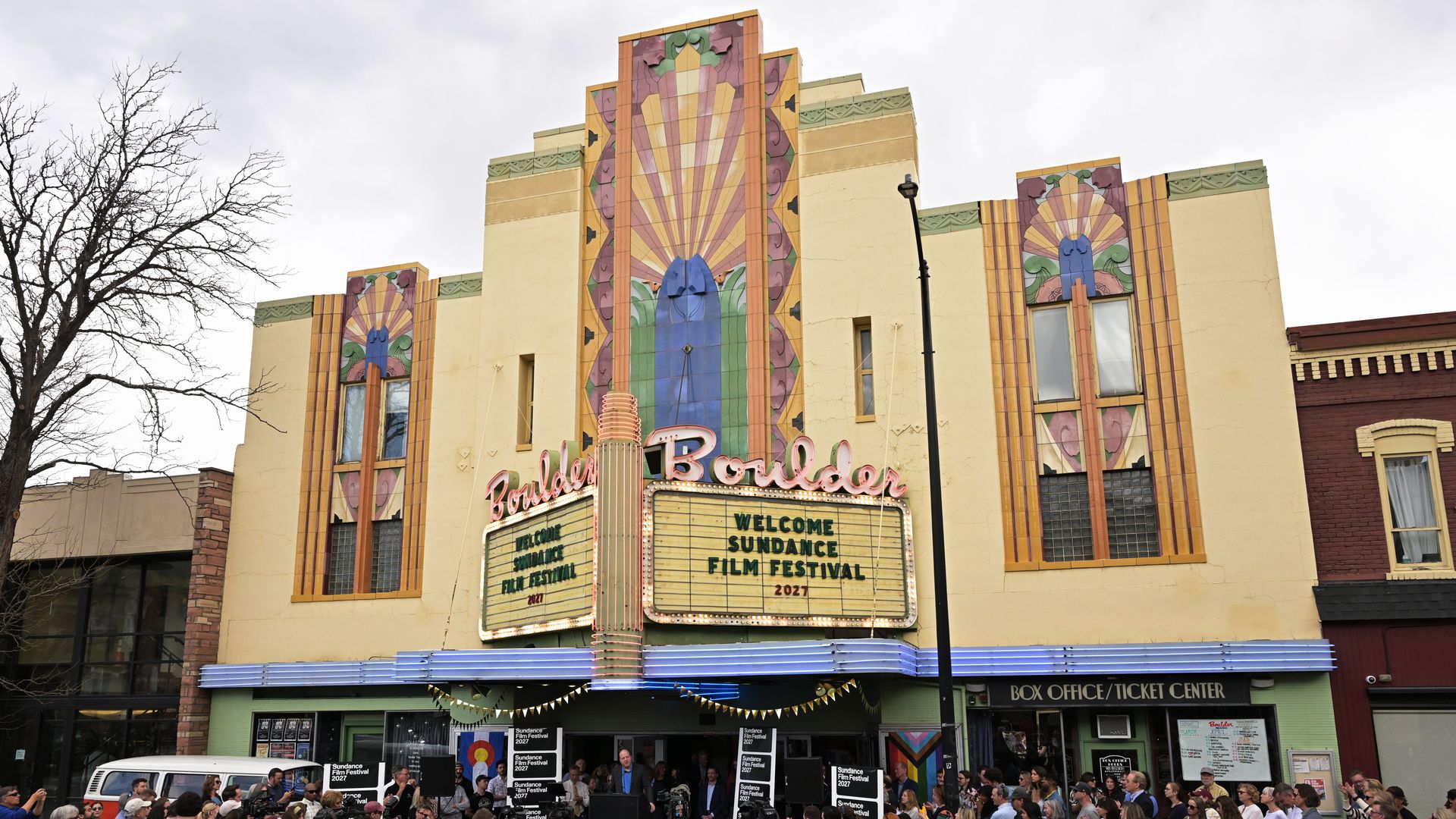 Art Deco theater with beige walls, orange-green trim, and pastel sunburst tile panels; a blue central figure dominates the facade. Marquee reads "Sundance Film Festival 2021". Crowds fill the street.