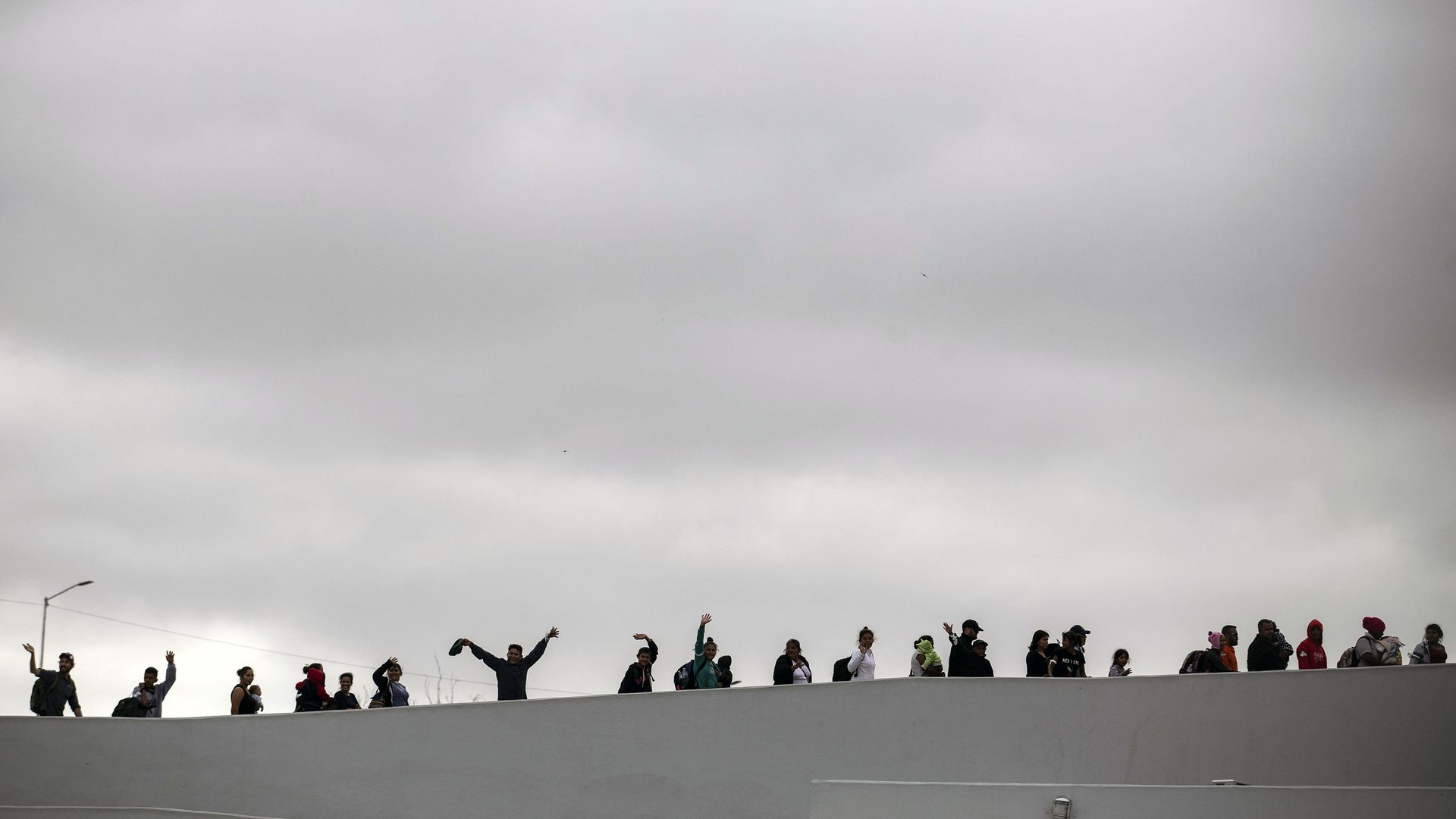 People waving overtop of a white wall with a grey sky in the background