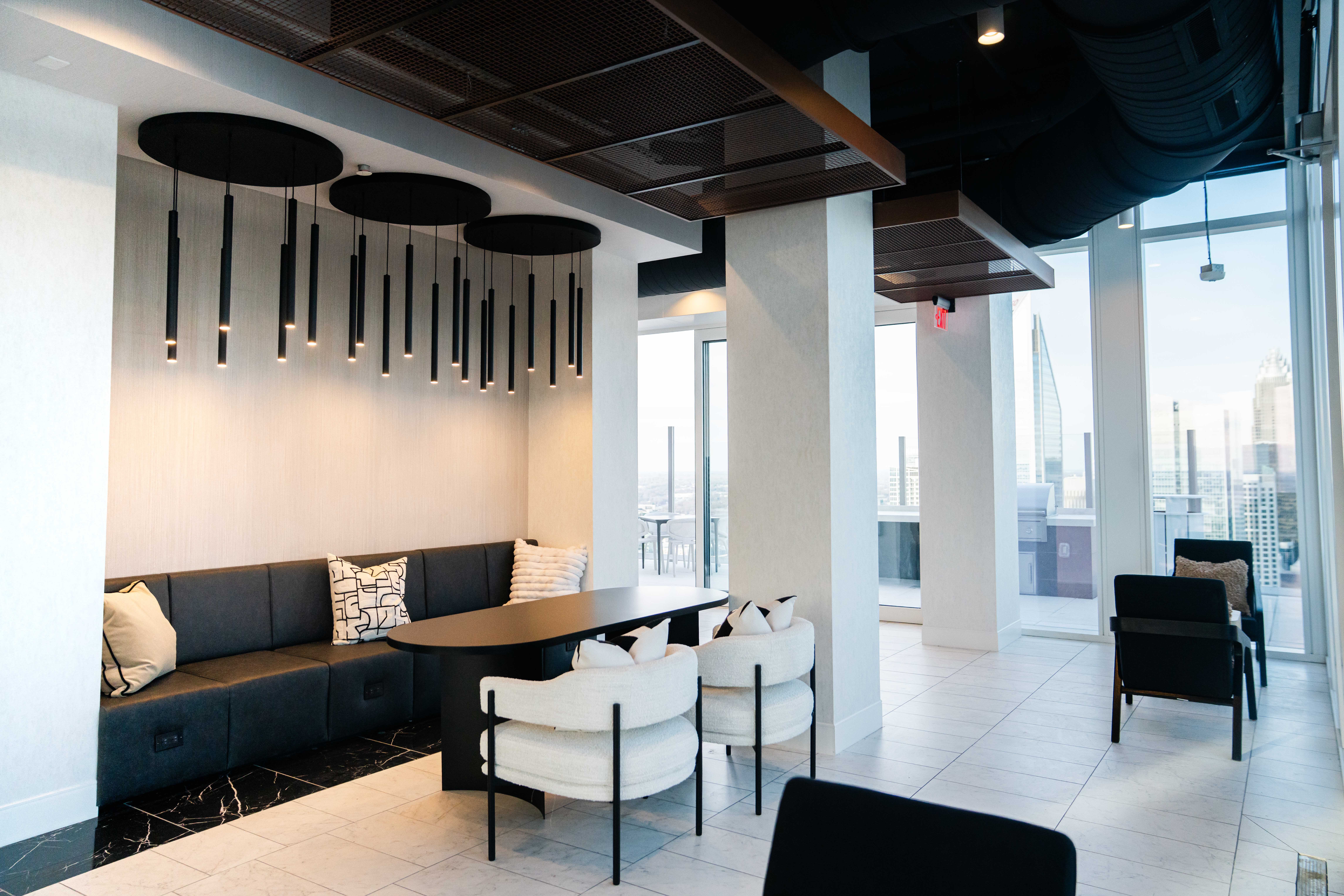 Modern office lounge with black cushioned bench, white chairs, pendant lights, large windows showcasing city skyline, and white tiled floor.