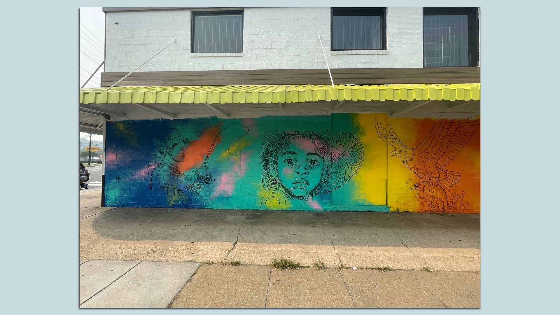 Colorful mural on a storefront wall under a yellow awning, featuring a blue-to-orange gradient background. Left: a fisherman; center: a girl with braided hair; right: a large bird of prey with a fish.