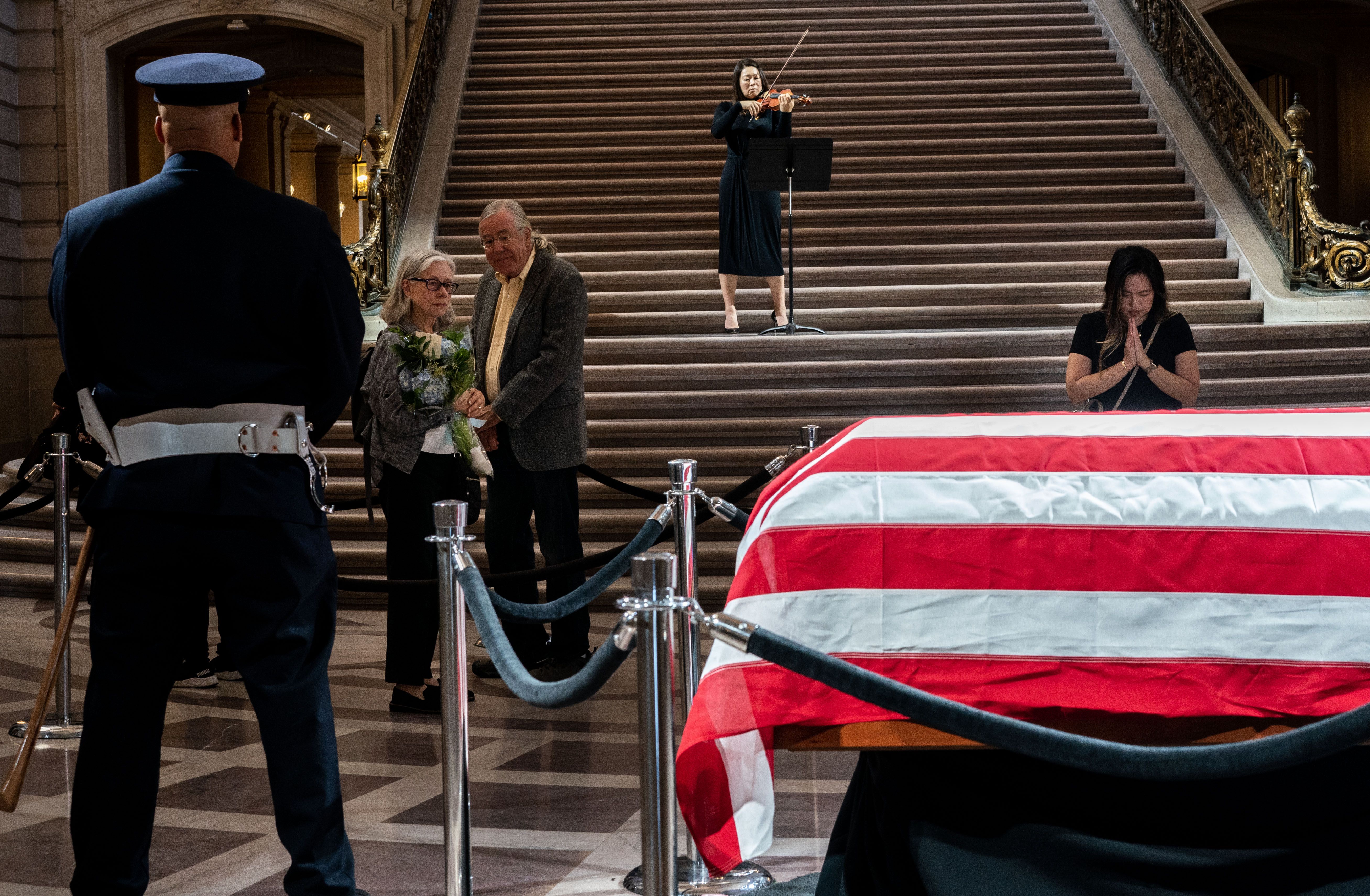 A mourner prays while someone plays the violin in the background on stairs. Feinstein's casket is in the foreground.
