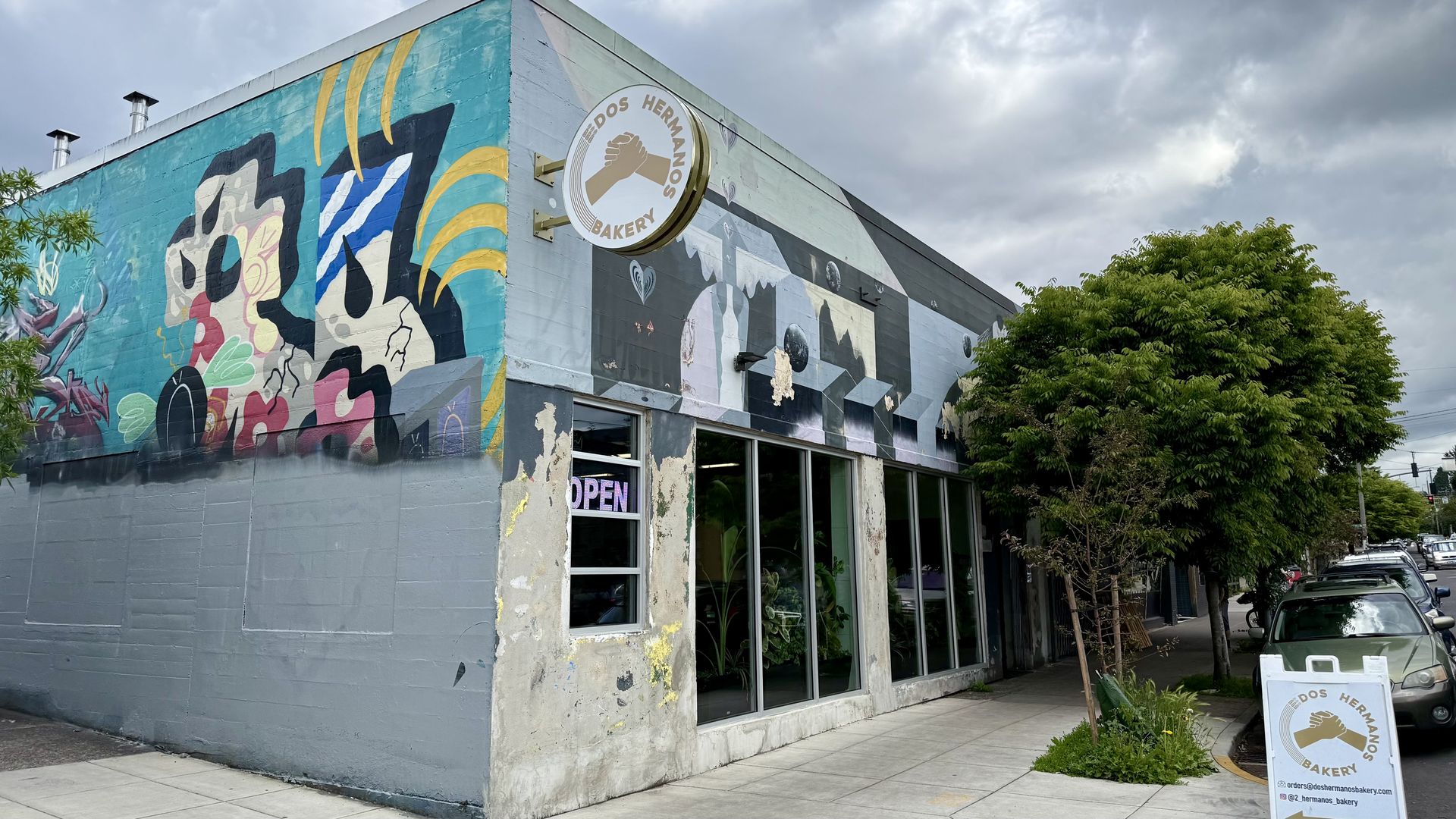Colorful mural-covered corner bakery building with a "Dos Hermanos Bakery" sign, open window, sidewalk trees, and a sandwich board on a cloudy day.