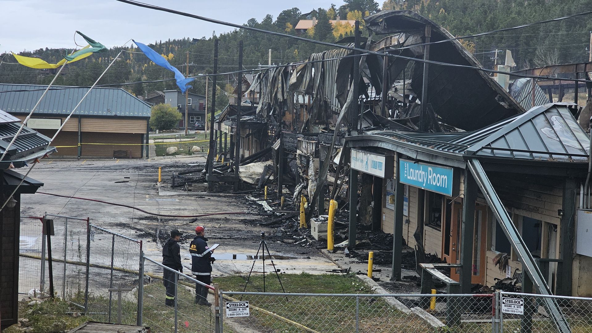 Burned-down building with significant fire damage, including a collapsed roof. Two investigators in black jackets stand in front of the site near a fence. A sign reads "The Laundry Room."