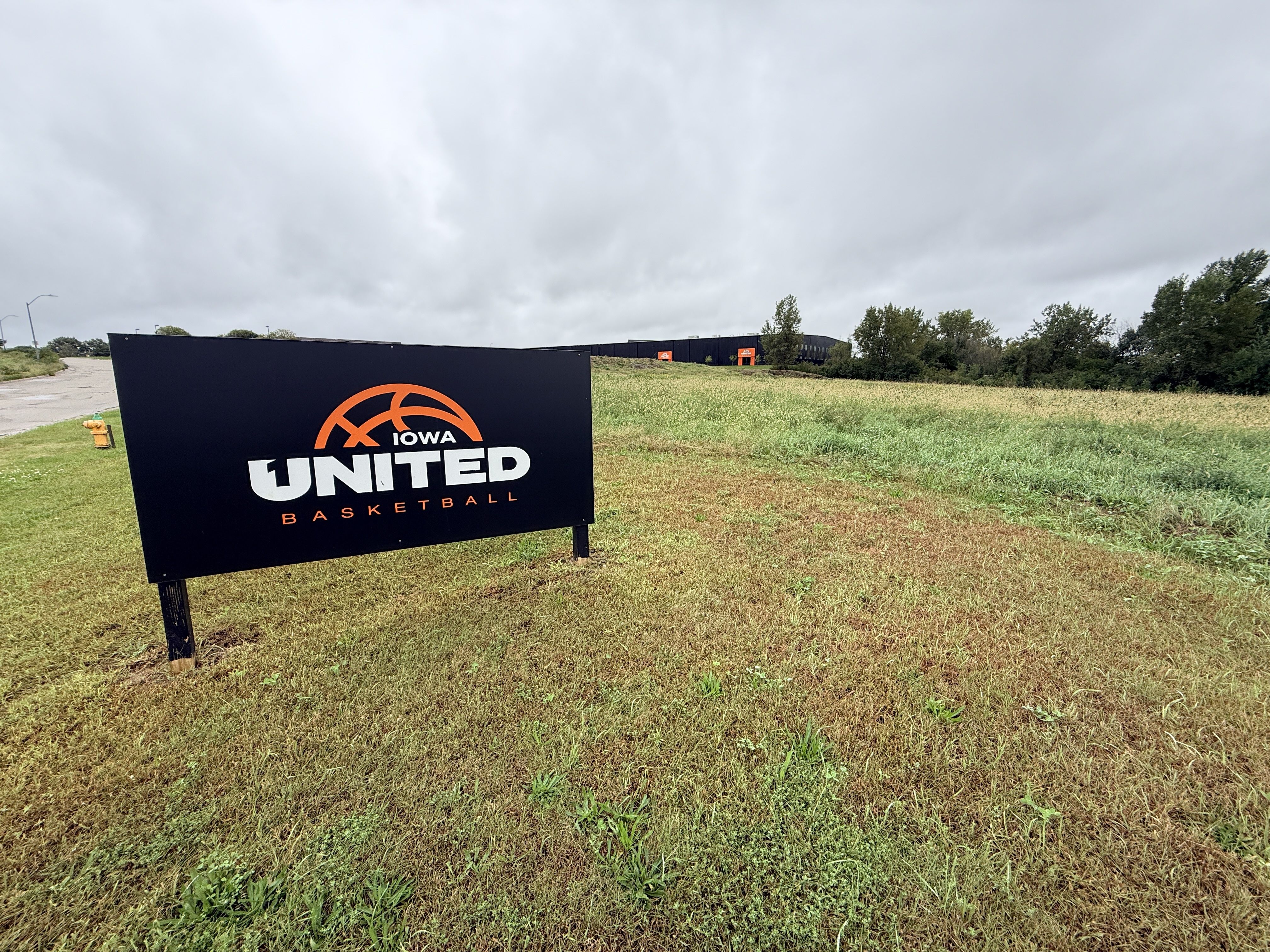 Overcast outdoor scene with a large black sign in the foreground reading "IOWA UNITED BASKETBALL" and an orange basketball logo; a long dark building, grassy field, and trees in the distance.