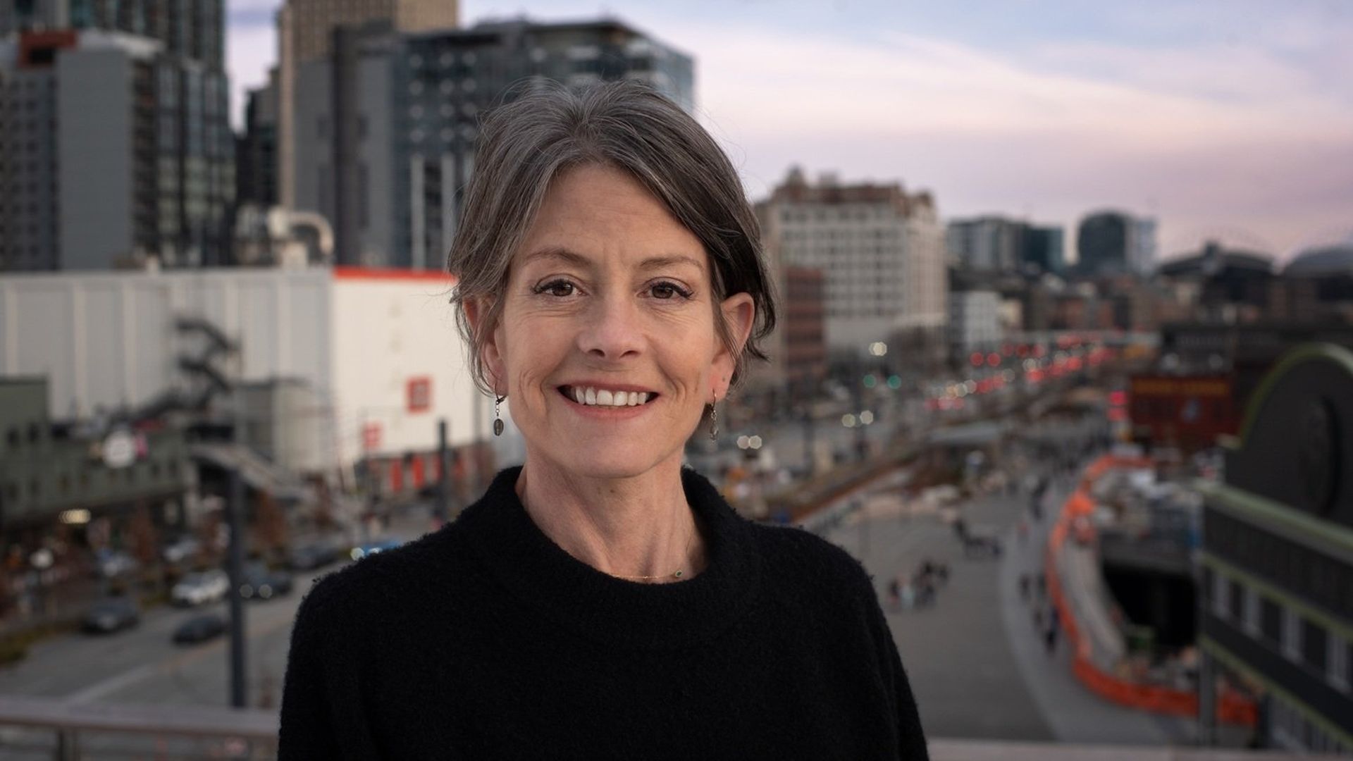 Smiling woman with short gray hair in a black sweater stands on a balcony, with a city skyline and busy street behind her at dusk.