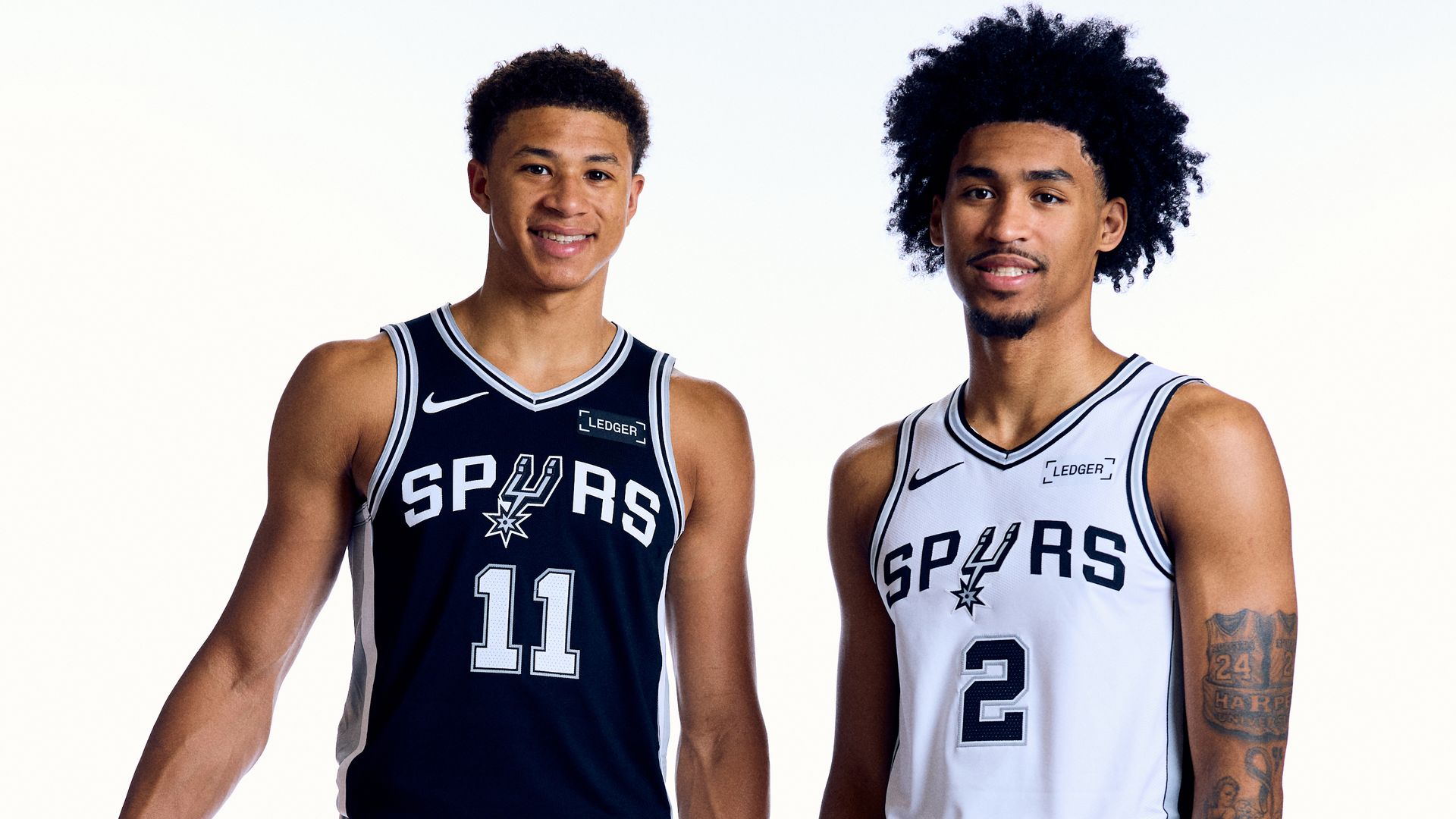 Carter Bryant (left) and Dylan Harper (left) pose shoulder to shoulder in front of a white background while wearing Spurs uniforms. 