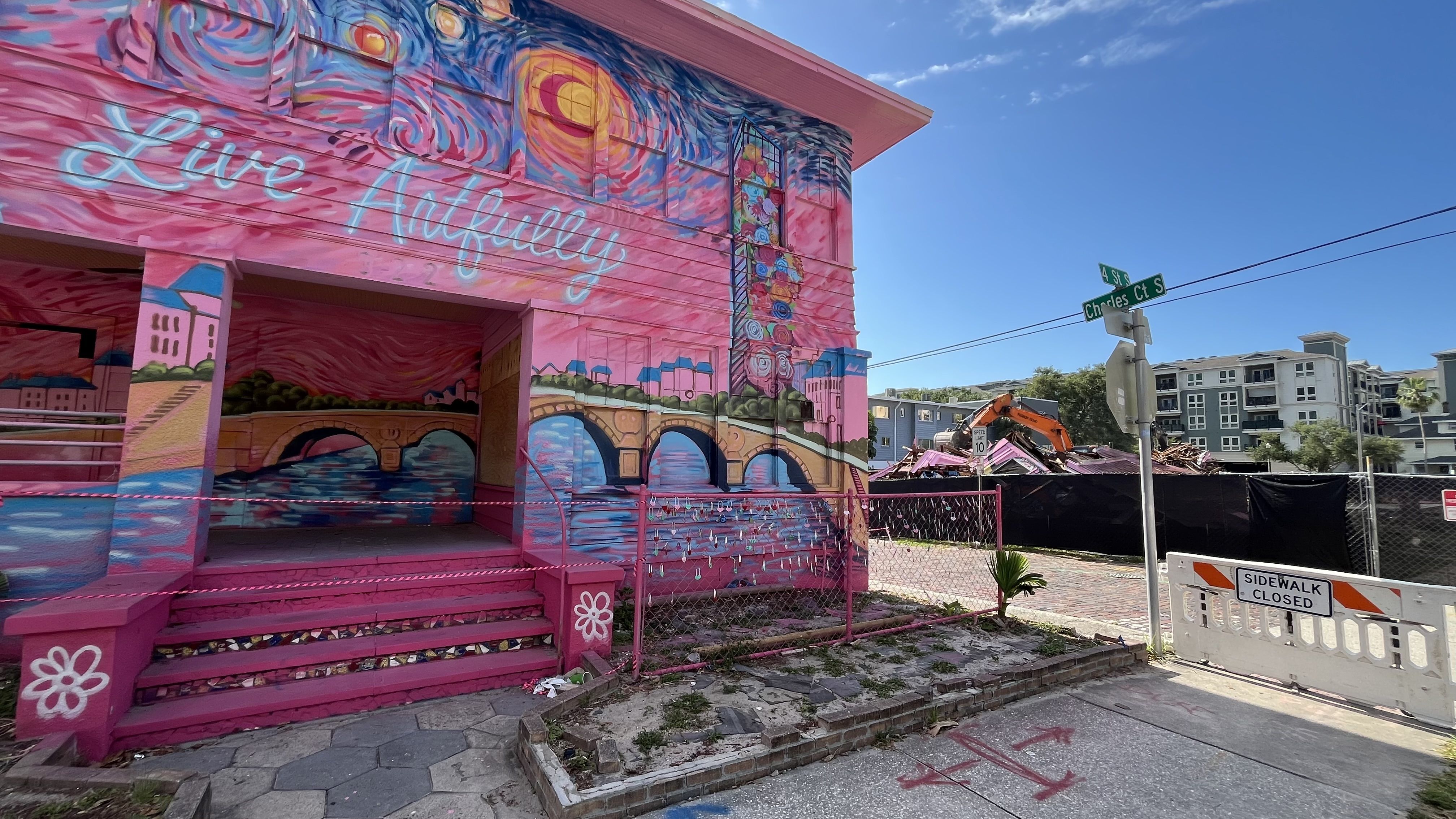 Bright pink mural building with blue cursive text Live Artfully, swirls of color, a bridge and roses. An orange excavator, street signs, and a sidewalk closed barrier on a sunny day.