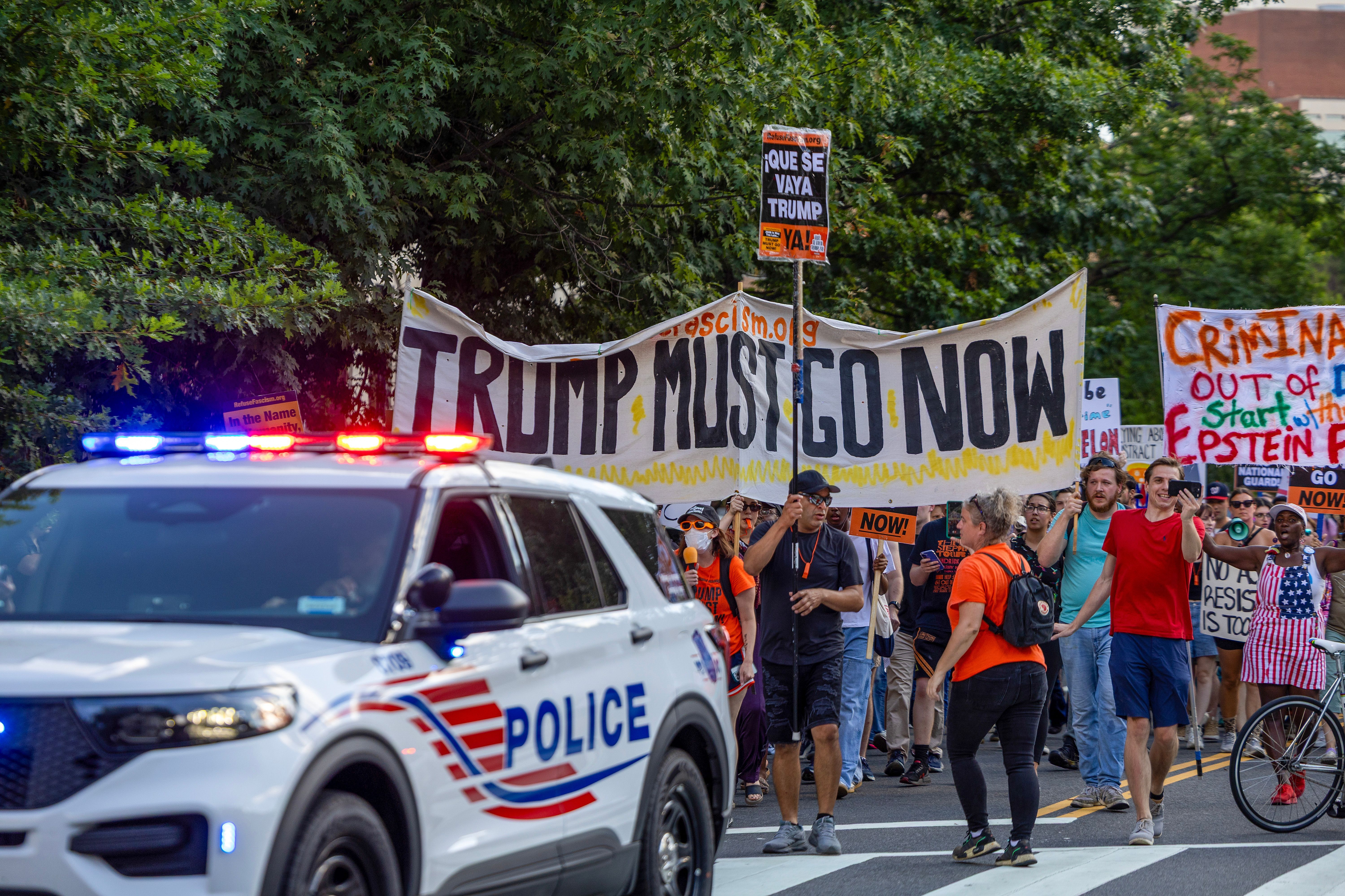 Protesters march with banners and signs reading "TRUMP MUST GO NOW" and "QUE SE VAYA TRUMP YA!" as a police SUV with flashing lights leads the crowd on a tree-lined street.