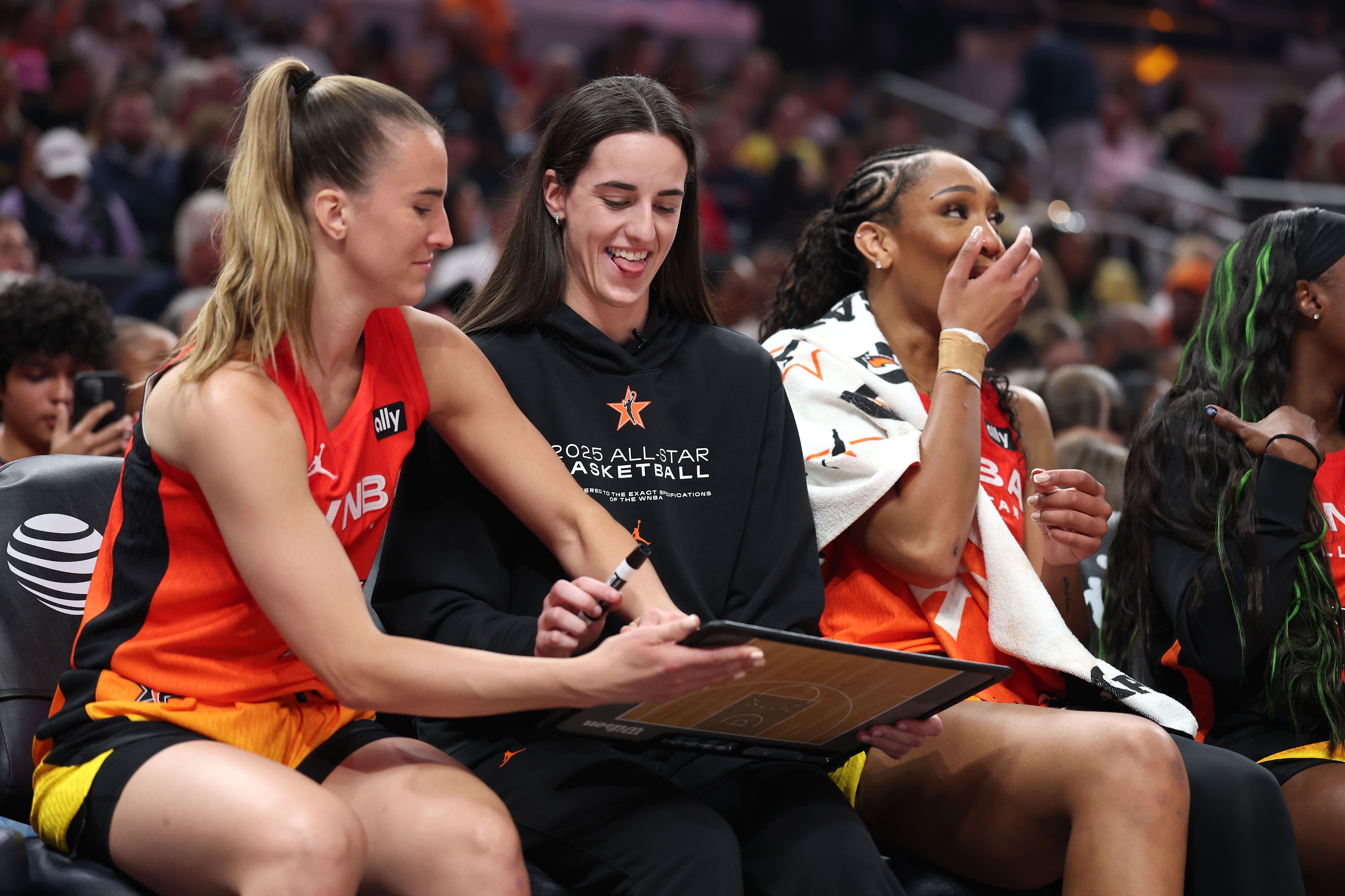 Three women seated courtside, two in orange basketball uniforms and one in a black 2025 All-Star Basketball hoodie, smiling and interacting with a clipboard with a basketball court diagram.