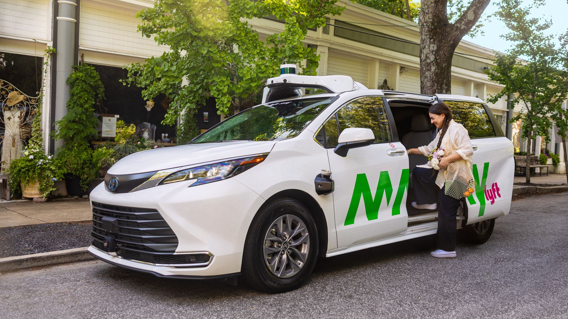 A woman carrying flowers and groceries enters a white autonomous minivan with the Lyft and May Mobility logos, on a tree-lined street.