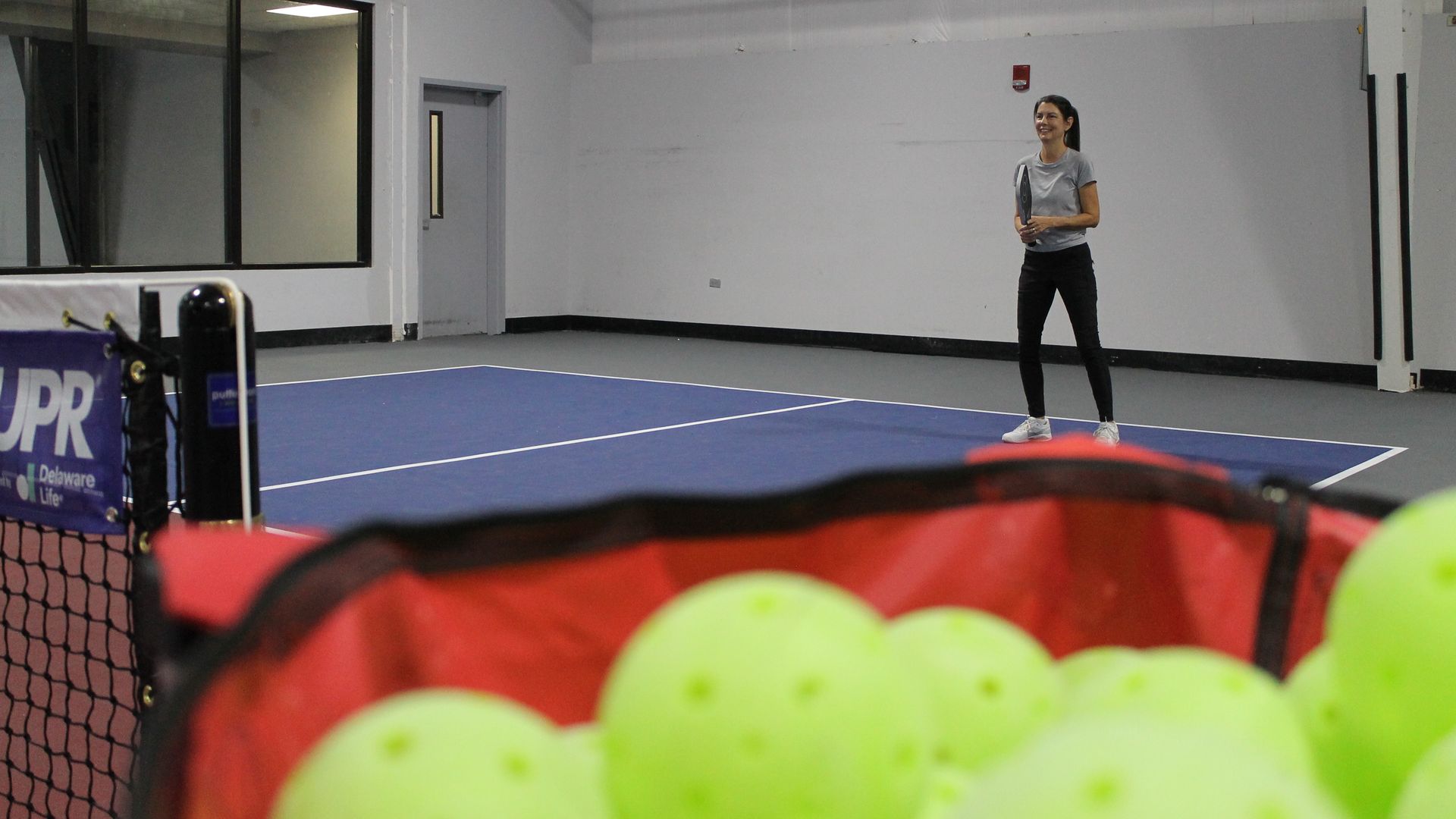 Woman in a gray shirt and black pants holding a pickleball paddle on an indoor blue pickleball court with a red basket of yellow pickleballs in the foreground.