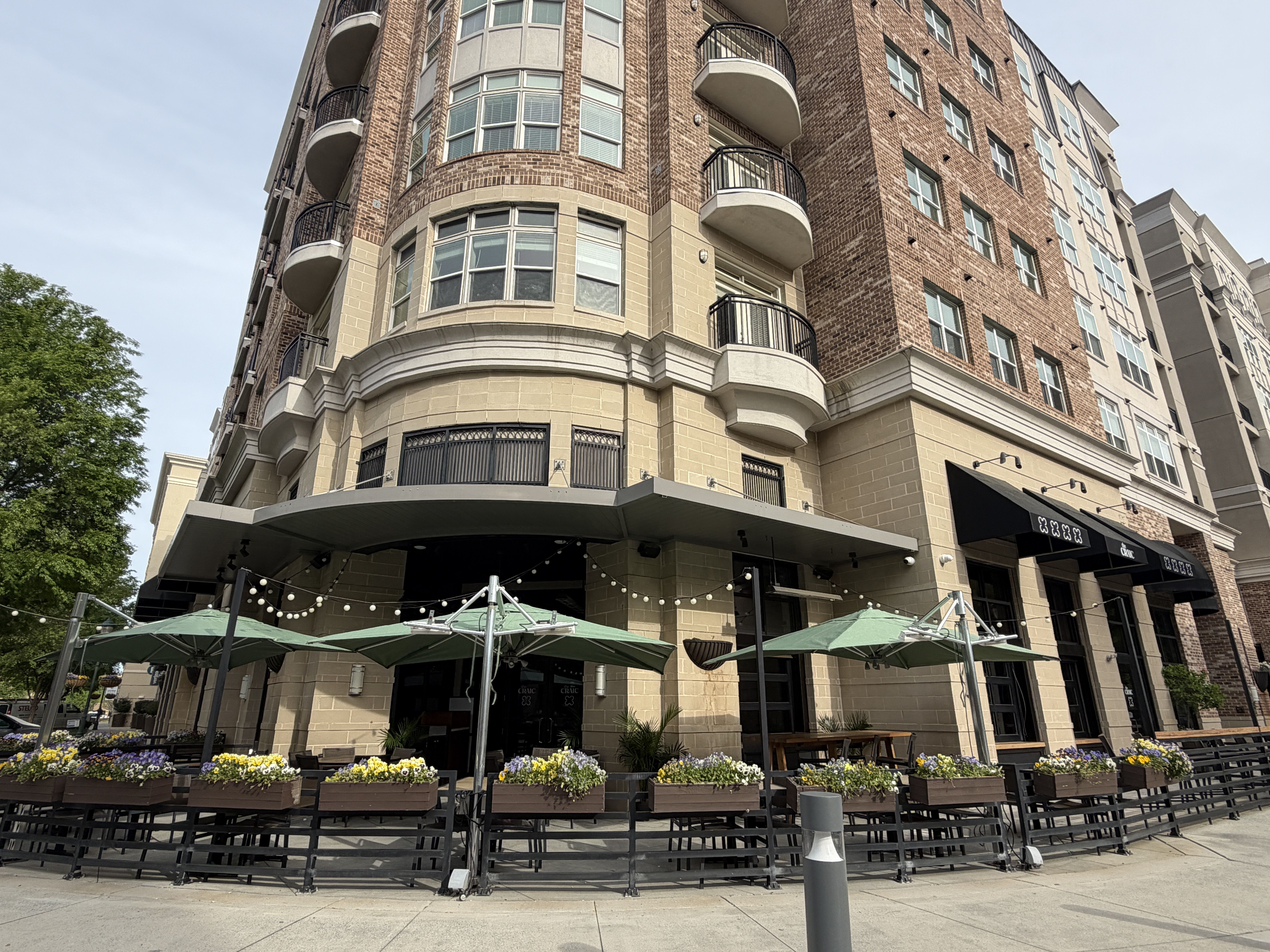 Multi-story brick building with curved balconies and beige stone base. Ground-floor shops with black awnings, outdoor seating under green umbrellas, string lights, and flower planters along sidewalk.