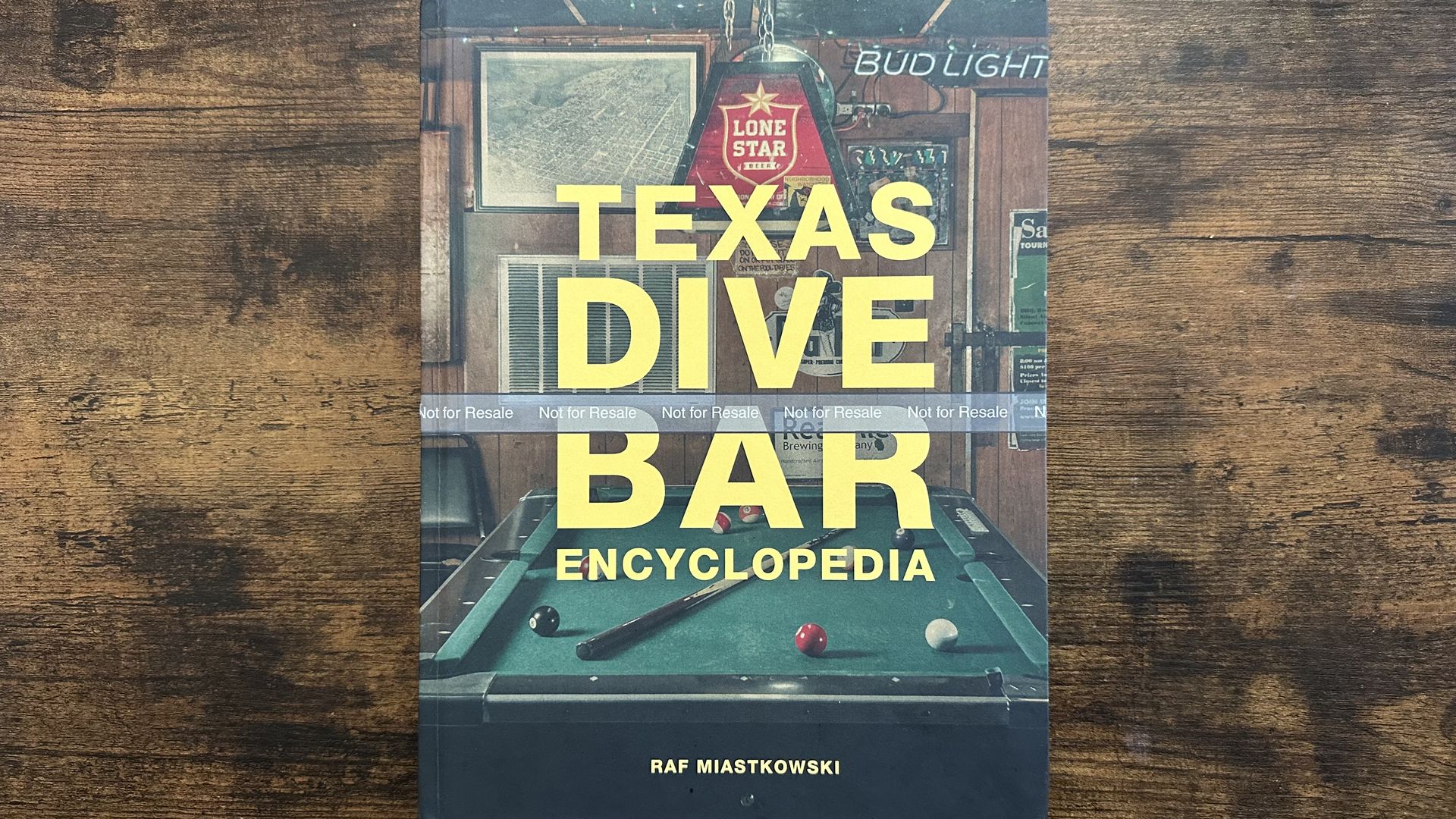 Book titled "Texas Dive Bar Encyclopedia" by Raf Miastkowski on a wooden surface with a pool table and bar signs visible on the cover, including a red Lone Star beer light.