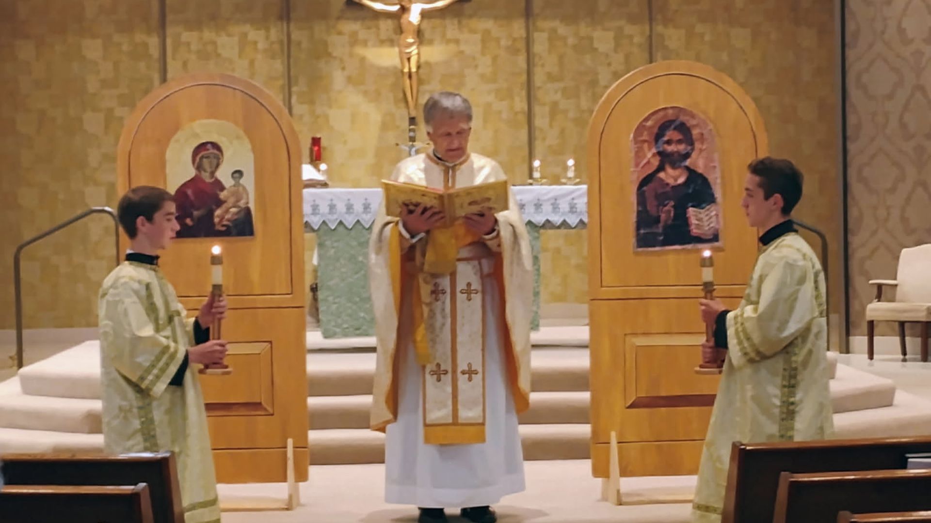 Priest in white and gold vestments reads from a book at church altar, flanked by two altar boys in gold robes holding candles, icons of Mary and Jesus behind them.