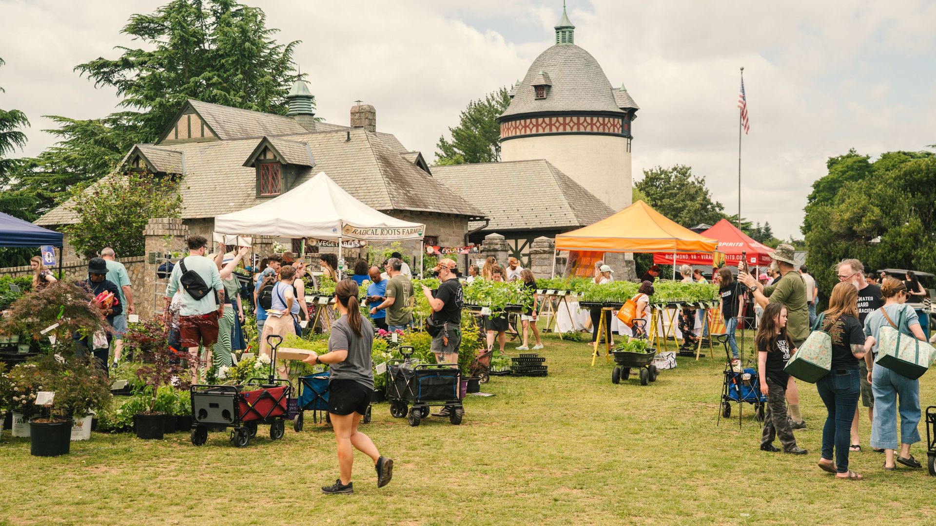 A lively outdoor market with people browsing plants under white, orange, blue tents near a stone building with a round turret and flagpole; trees and a cloudy sky above.