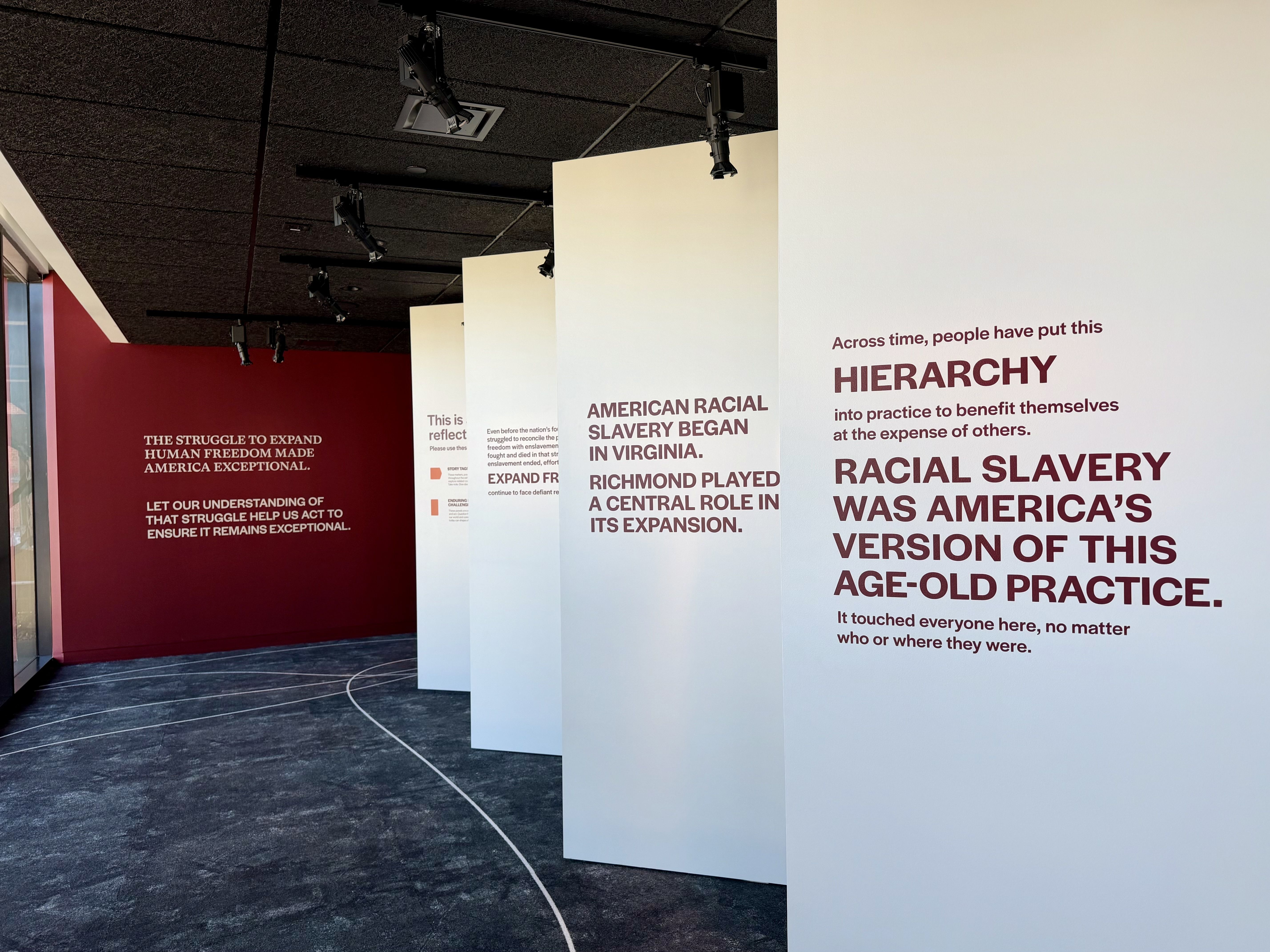 Exhibition gallery with tall white panels bearing bold maroon text about hierarchy and racial slavery; maroon wall on left with white text; track lighting, curved white line on dark floor.