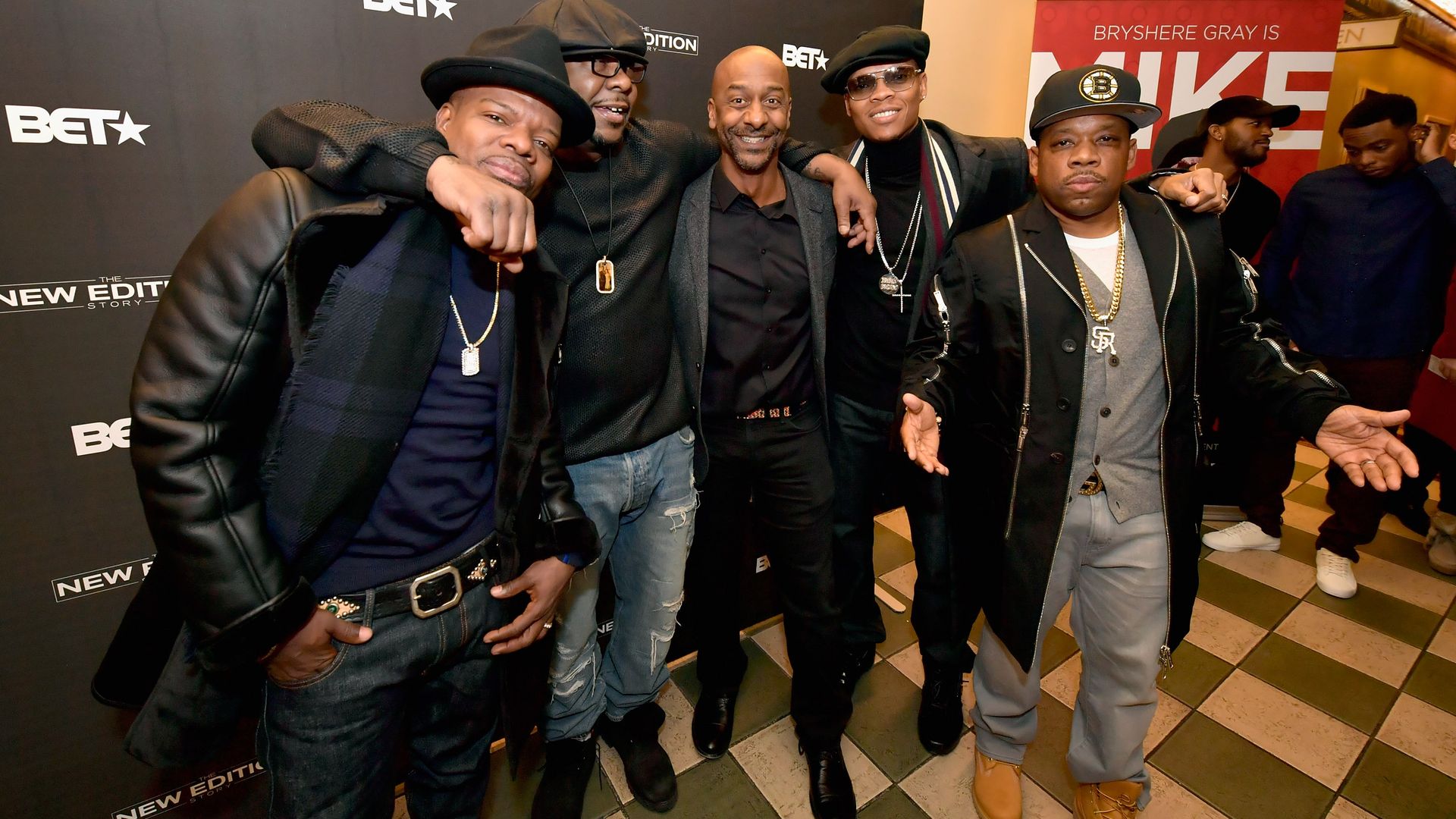 Five men posing together at a BET event for "The New Edition Story," dressed in dark and casual outfits with accessories and hats, smiling against a black branded backdrop.