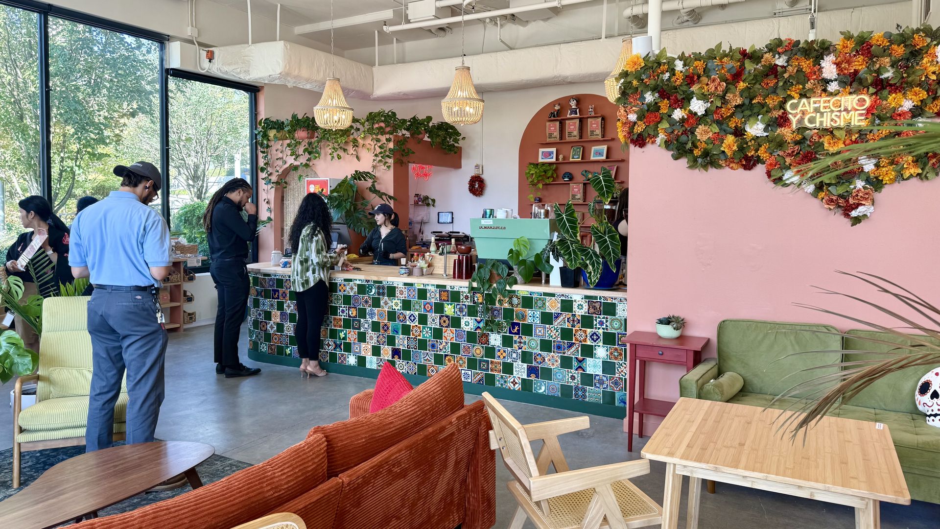 Interior of a colorful coffee shop with mosaic tile counter, pink walls, flower decor, green and orange furniture, large windows, and customers ordering at the counter.