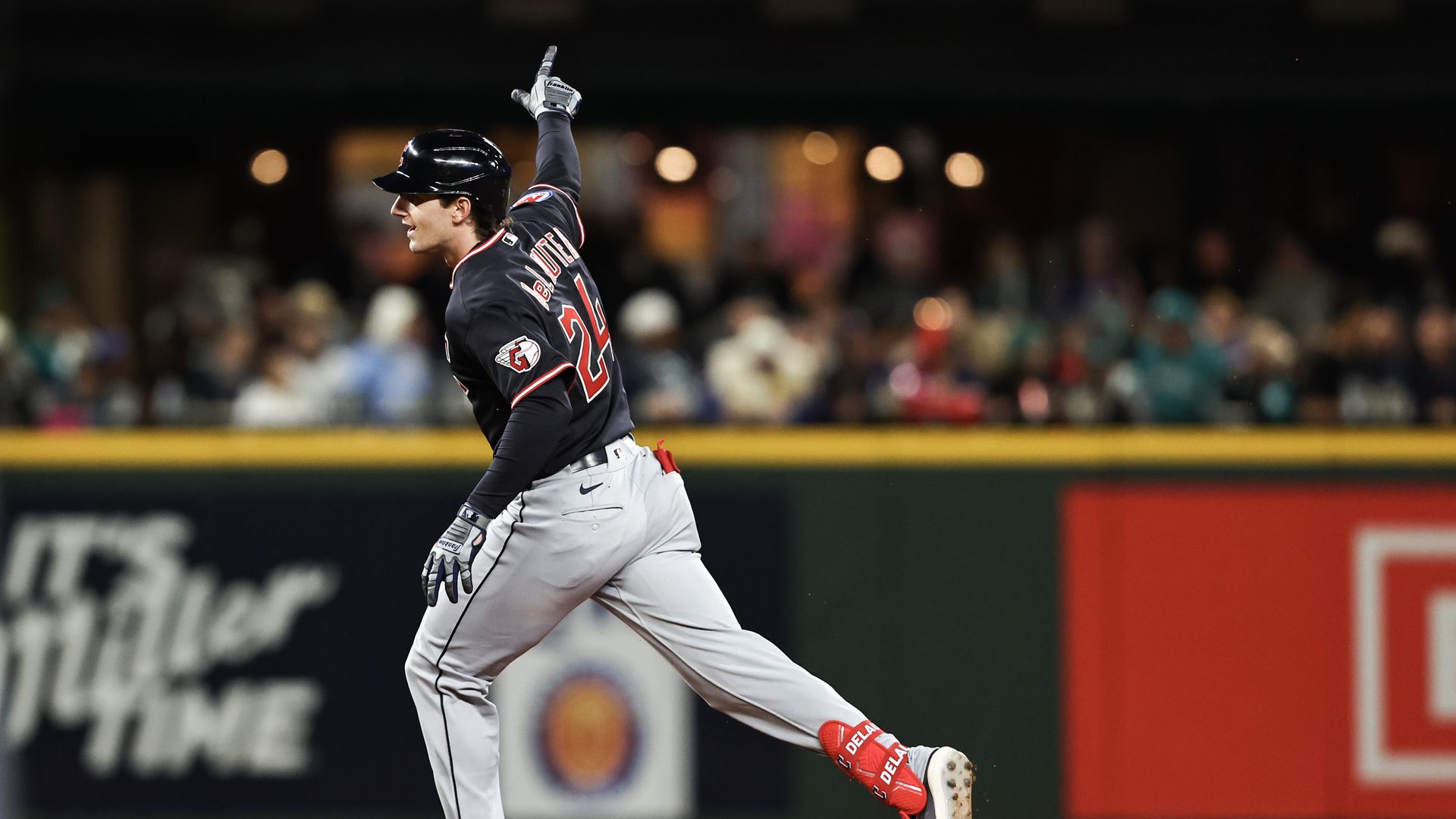 Baseball player in a black jersey with red trim and gray pants, mid-trot on dirt, raising one gloved hand and pointing a finger up as the crowd watches from the blurred stands.