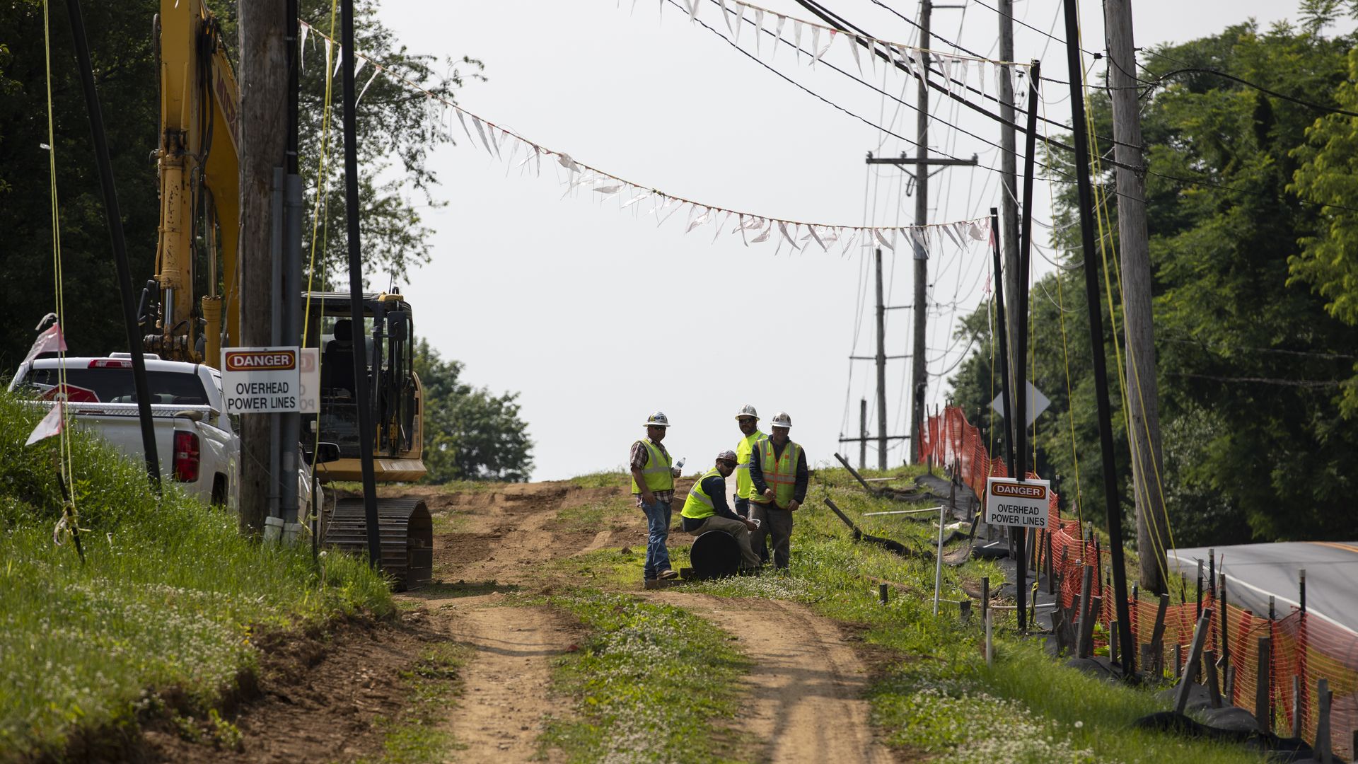 In this image, construction workers stand on a dirt road near a paved road.